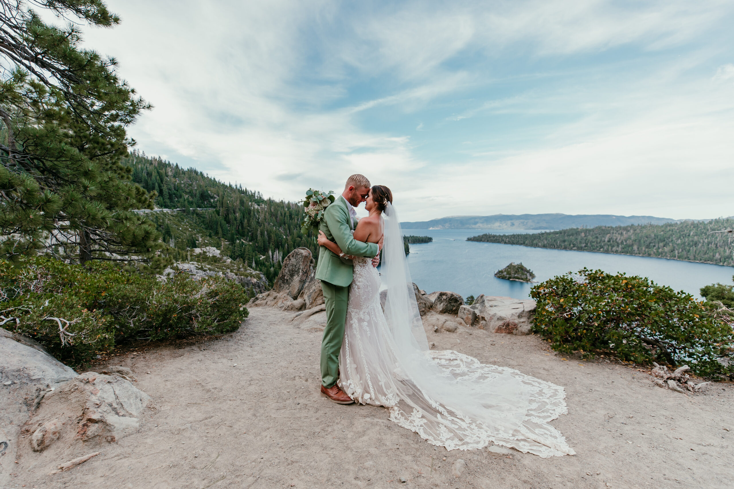 Bride and groom embracing during their Emerald Bay elopement in Lake Tahoe, with a sweeping view of Fannette Island and forested mountains behind them.