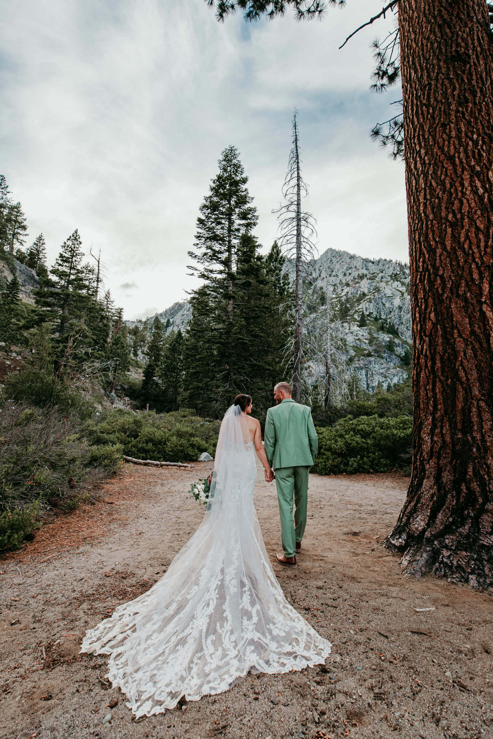 Bride and groom walking hand in hand through the forest after their Emerald Bay elopement, with mountain peaks rising in the background.