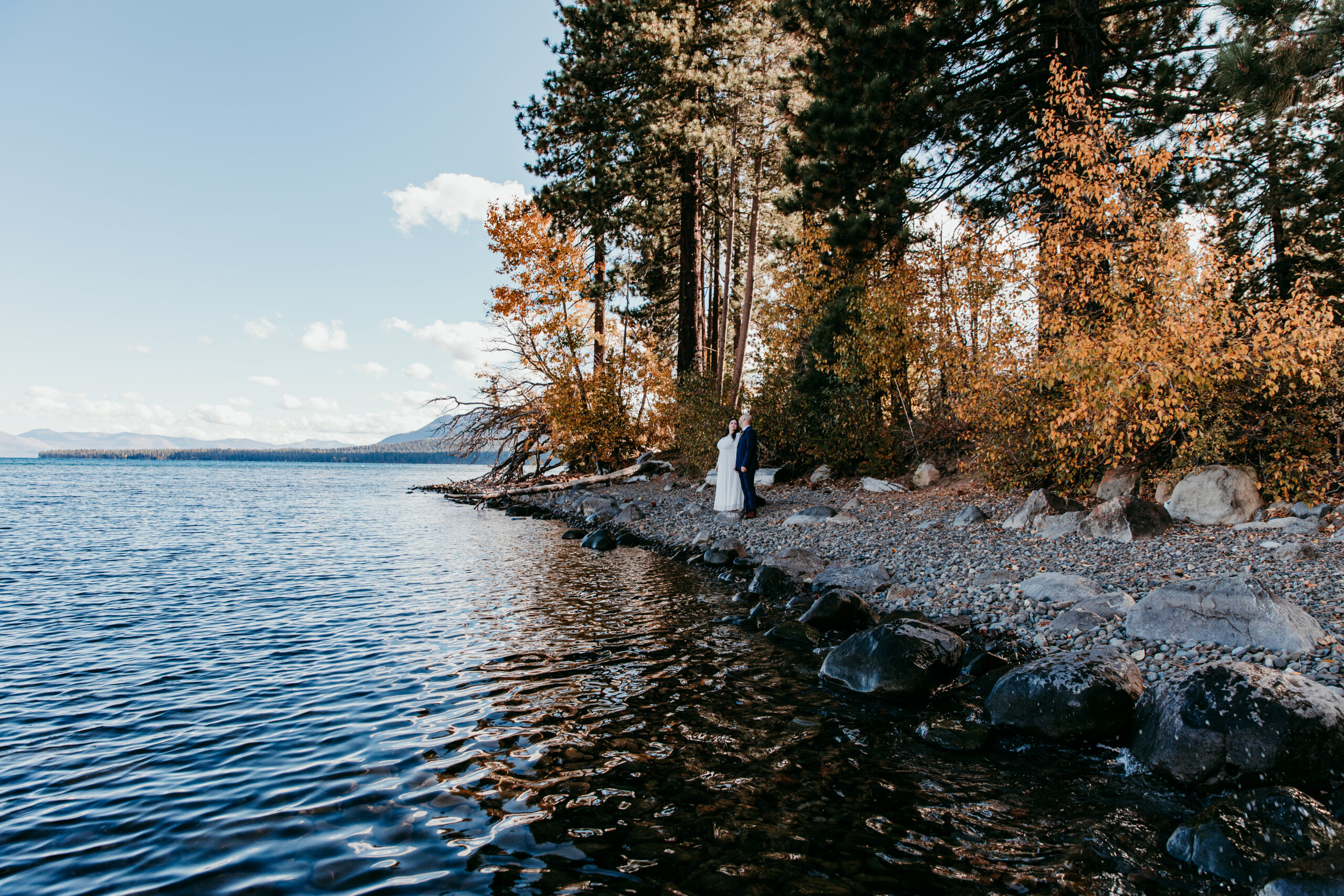 Couple standing by the shore of Lake Tahoe surrounded by golden fall trees, capturing the warm and peaceful autumn atmosphere that makes fall one of the best times to elope in Lake Tahoe.