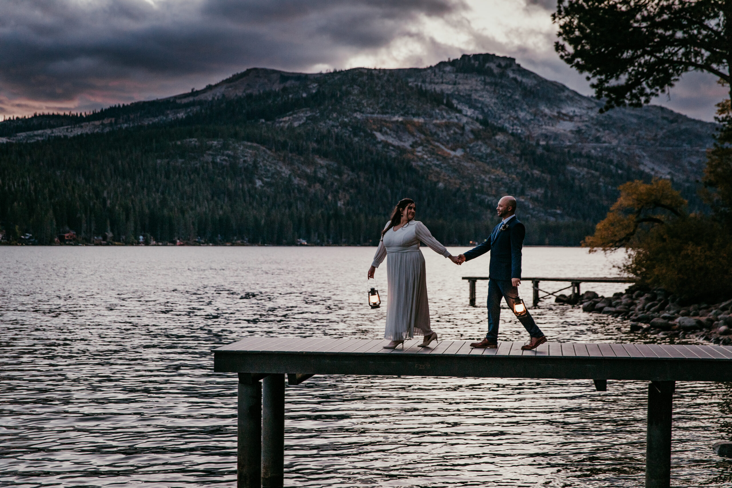 Bride and groom walking hand in hand on a lakeside dock with lanterns during a Lake Tahoe elopement, captured in moody lake tahoe elopement photos at dusk