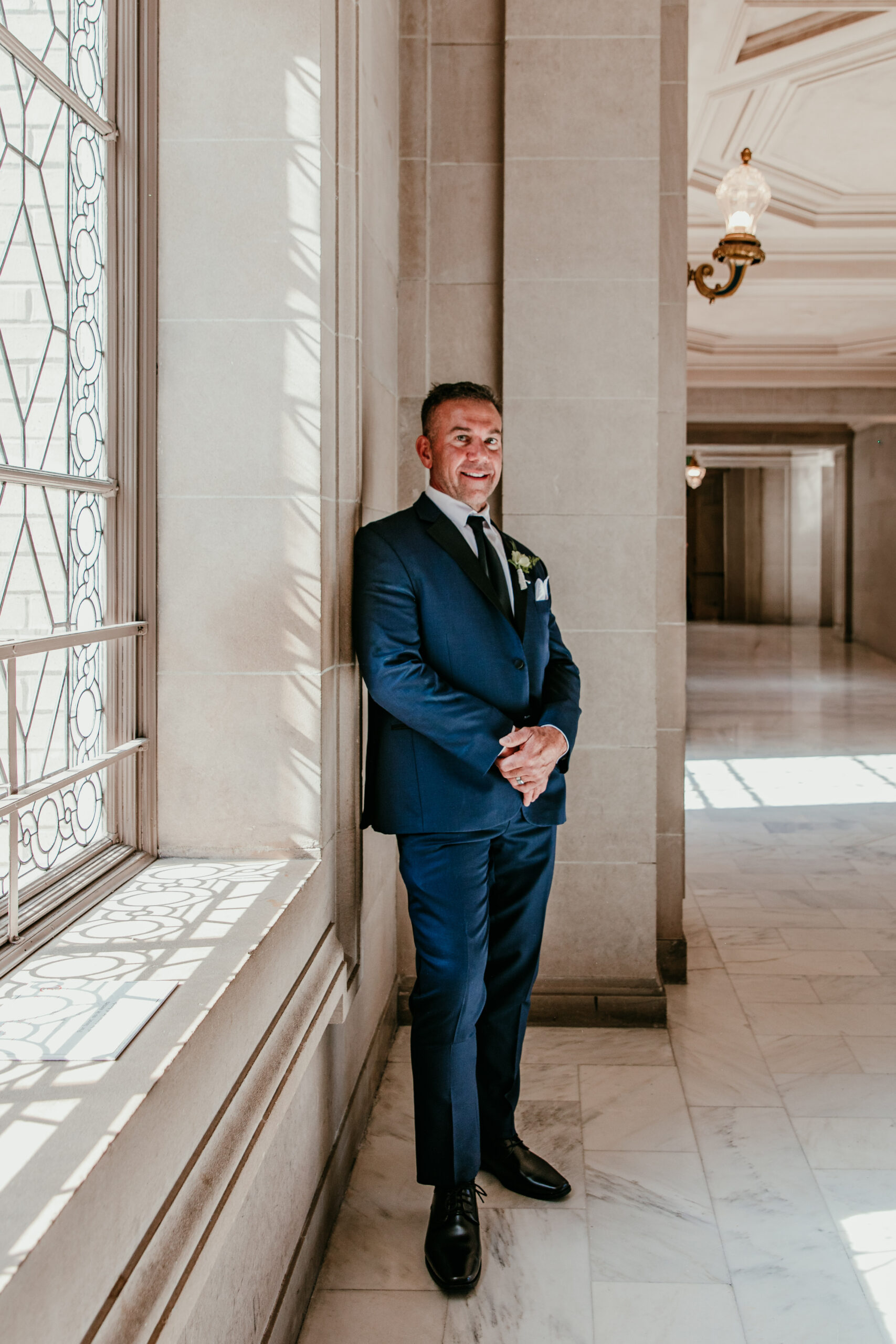 Groom standing by a sunlit window inside San Francisco City Hall during his elopement, photographed by a san francisco elopement photographer.