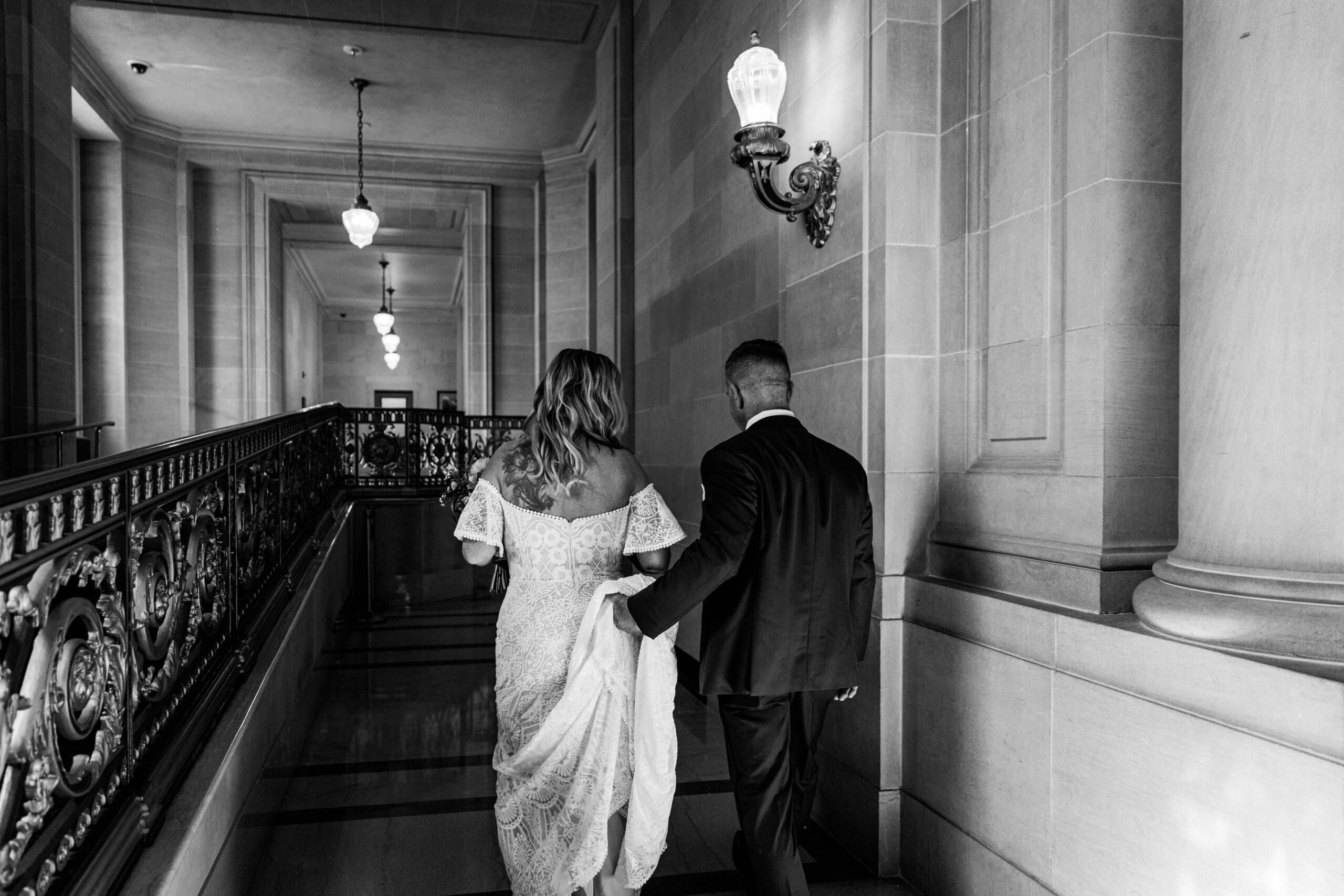 Black and white photo of the couple walking through the hallway inside San Francisco City Hall after their elopement ceremony, captured by a san francisco elopement photographer.
