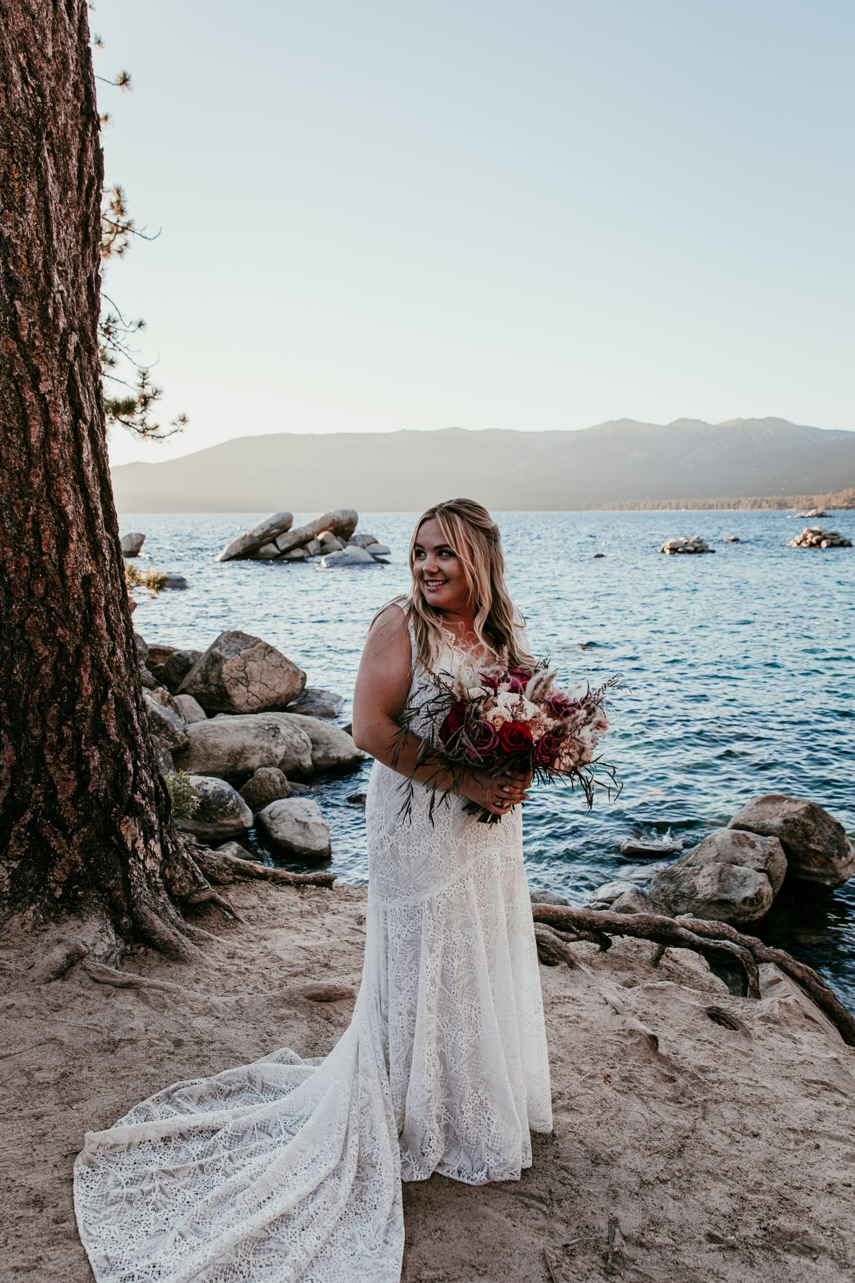 Bride holding a bouquet near the water at during a lake elopement, wearing a lace elopement dress with a soft flowing train