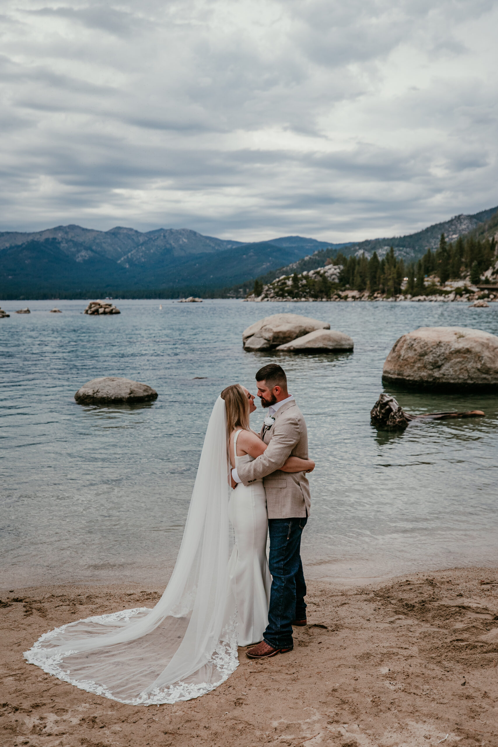 Couple embracing at the edge of a lake during a lakeside elopement, bride wearing a simple elopement dress with a long veil