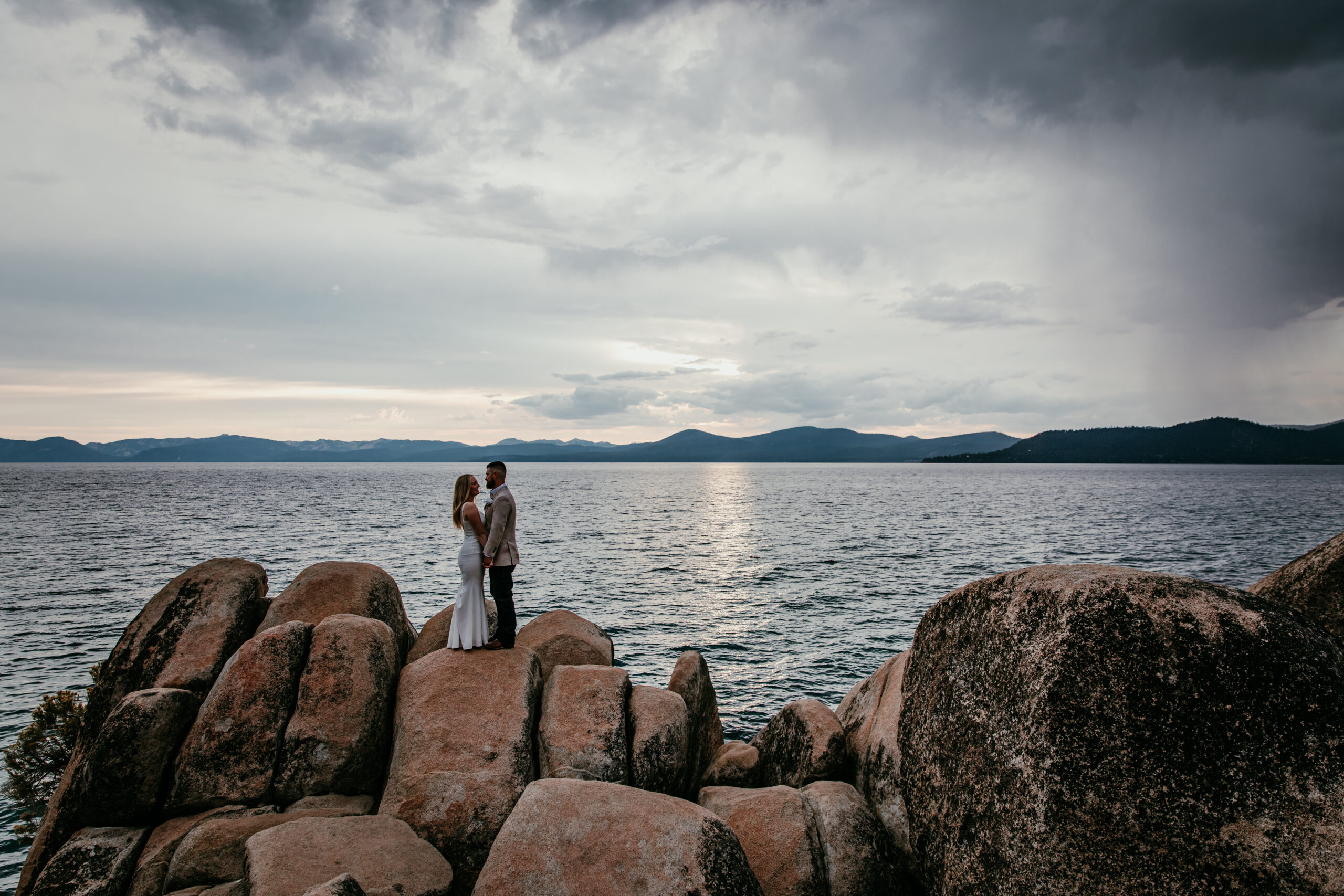 Couple standing together on large granite rocks during a Lake Tahoe California elopement, with the lake and mountain skyline under moody clouds.