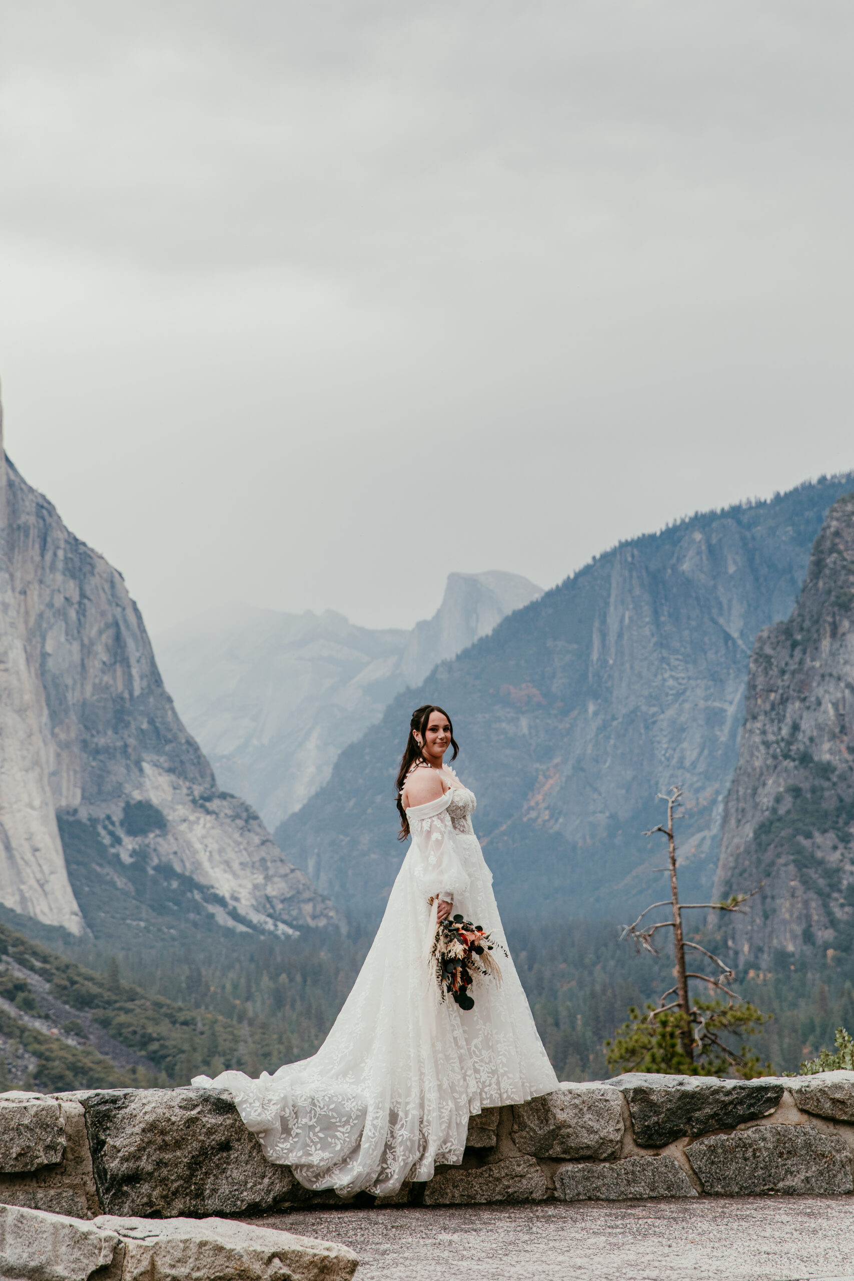 Bride standing at a scenic overlook in Yosemite during a mountain elopement, wearing an off-the-shoulder lace elopement dress