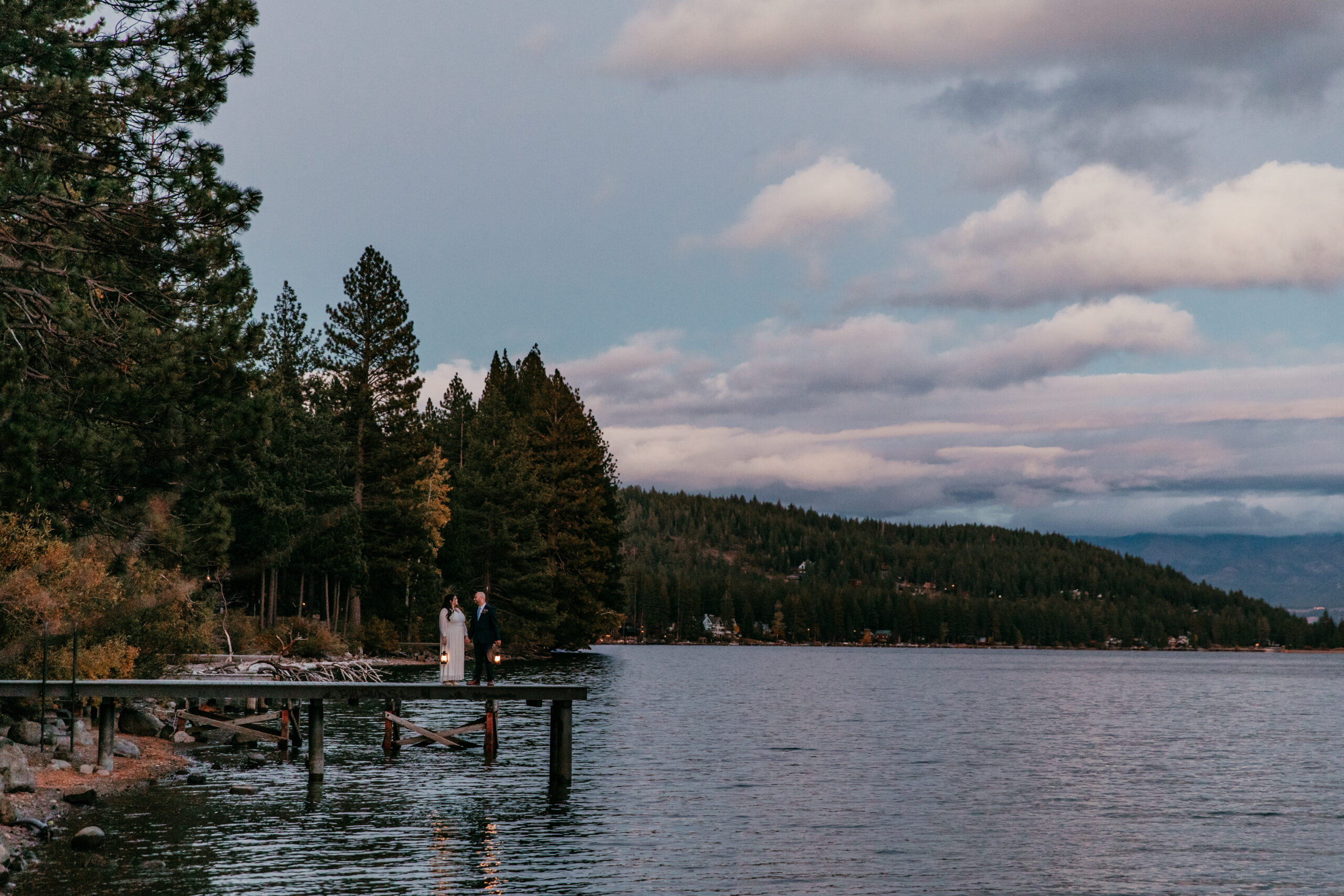 Couple getting married in Lake Tahoe standing on a lakeside dock at dusk with pine trees, calm water, and soft evening clouds.