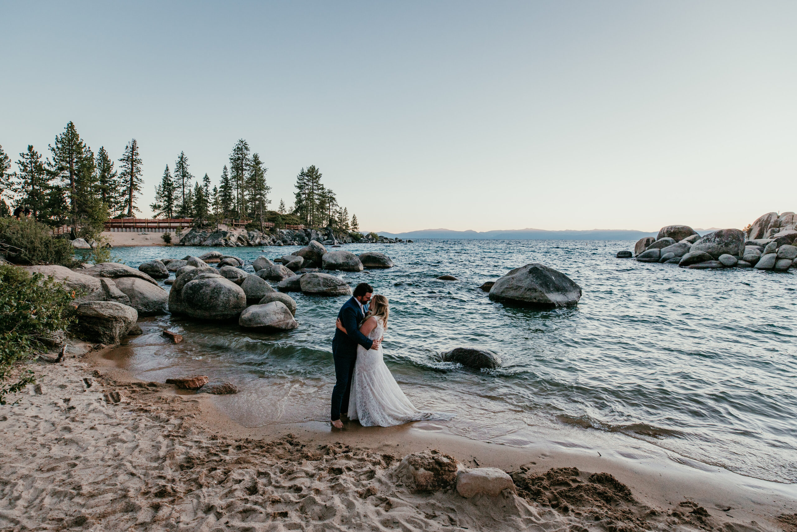 Couple getting married in Lake Tahoe standing barefoot at the water’s edge with granite boulders, pine trees, and calm lake views.