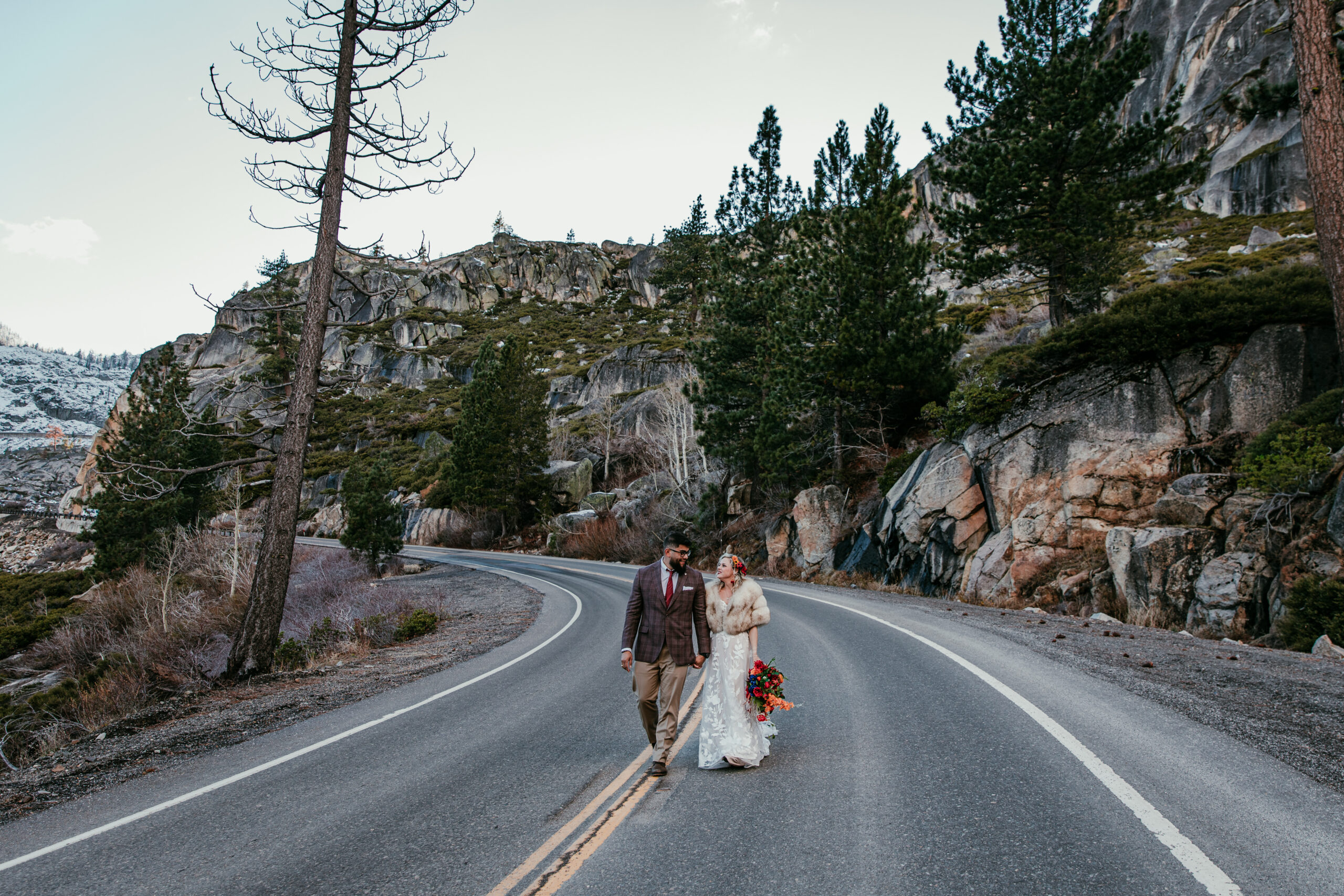 Couple walking together on a mountain road during an elopement, bride wearing a long lace elopement dress and fur wrap.