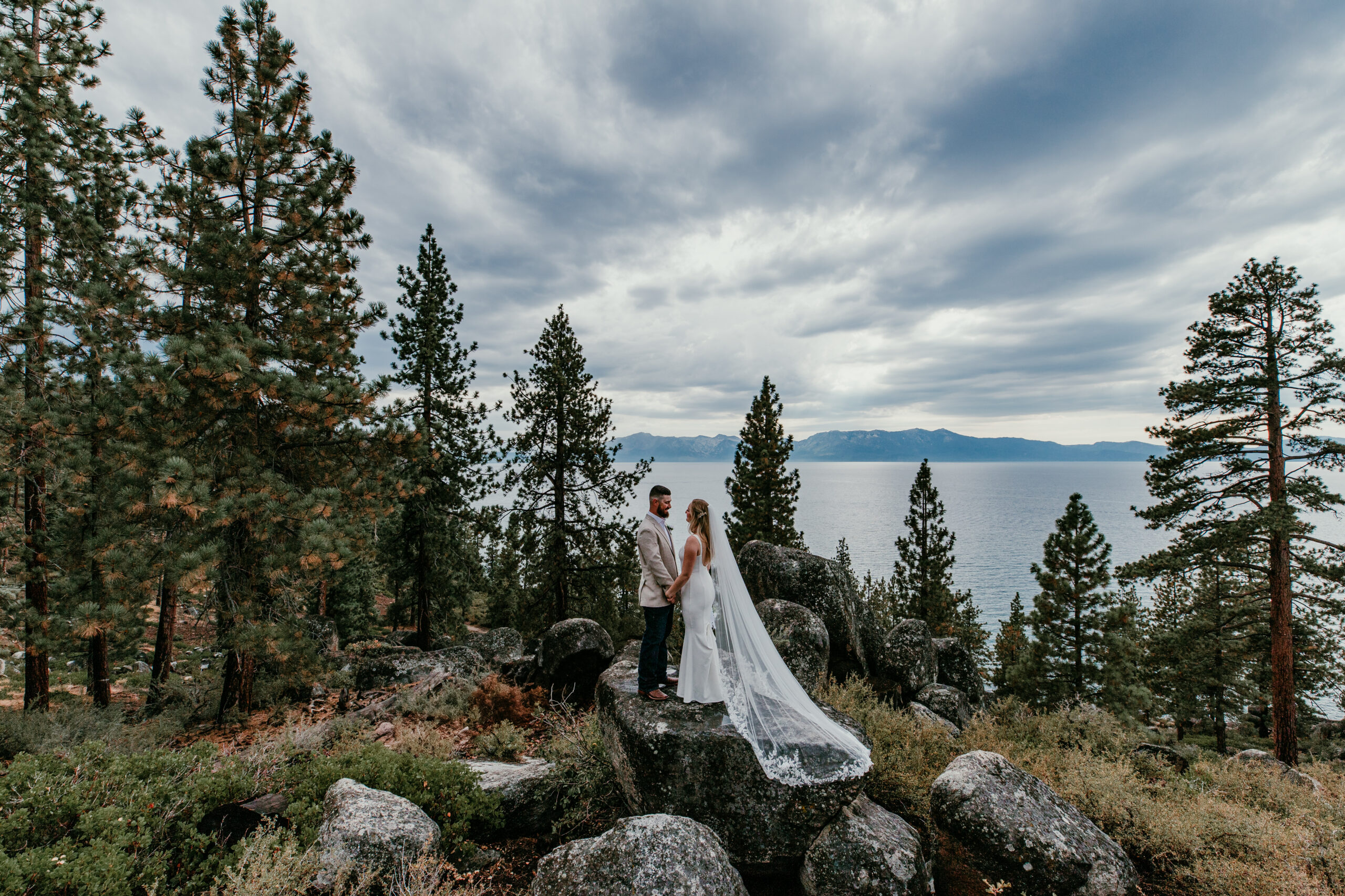 Wide view of Lake Tahoe elopement ceremony on rocky overlook with bride and groom surrounded by pine trees and mountain lake