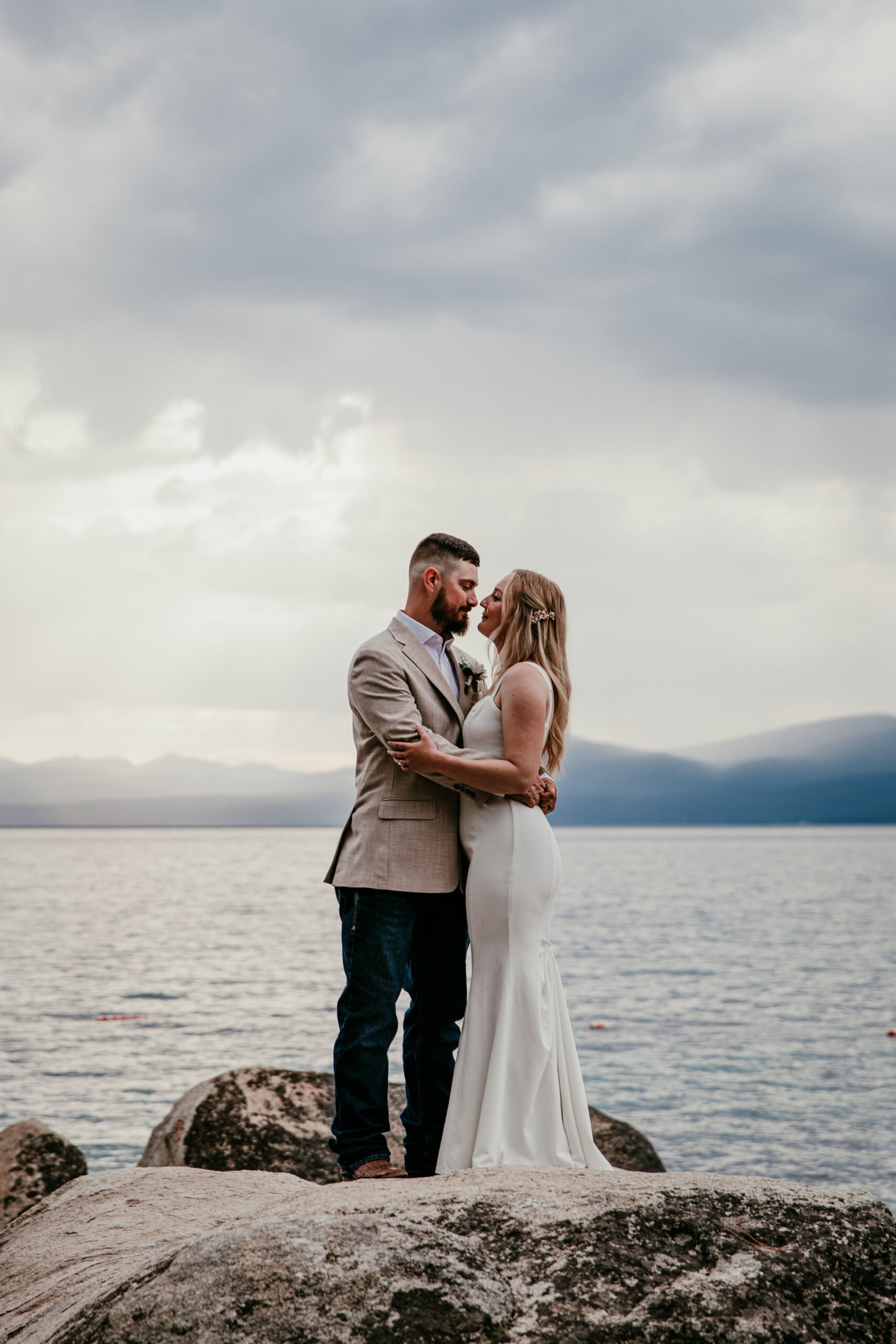 Intimate wedding couple embracing on rocks at Lake Tahoe with moody sky and mountain views