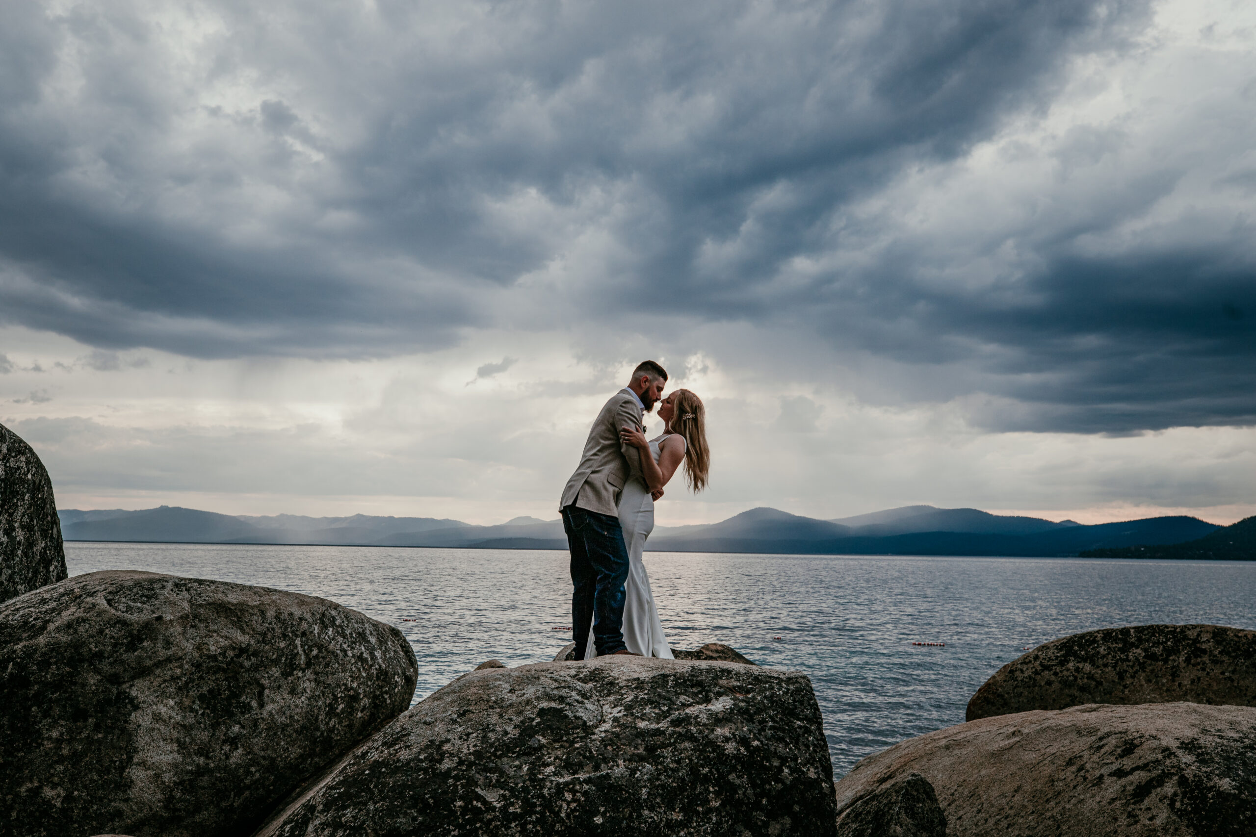 Couple kissing on lakeside rocks after their small wedding ceremony in Lake Tahoe with moody sky.