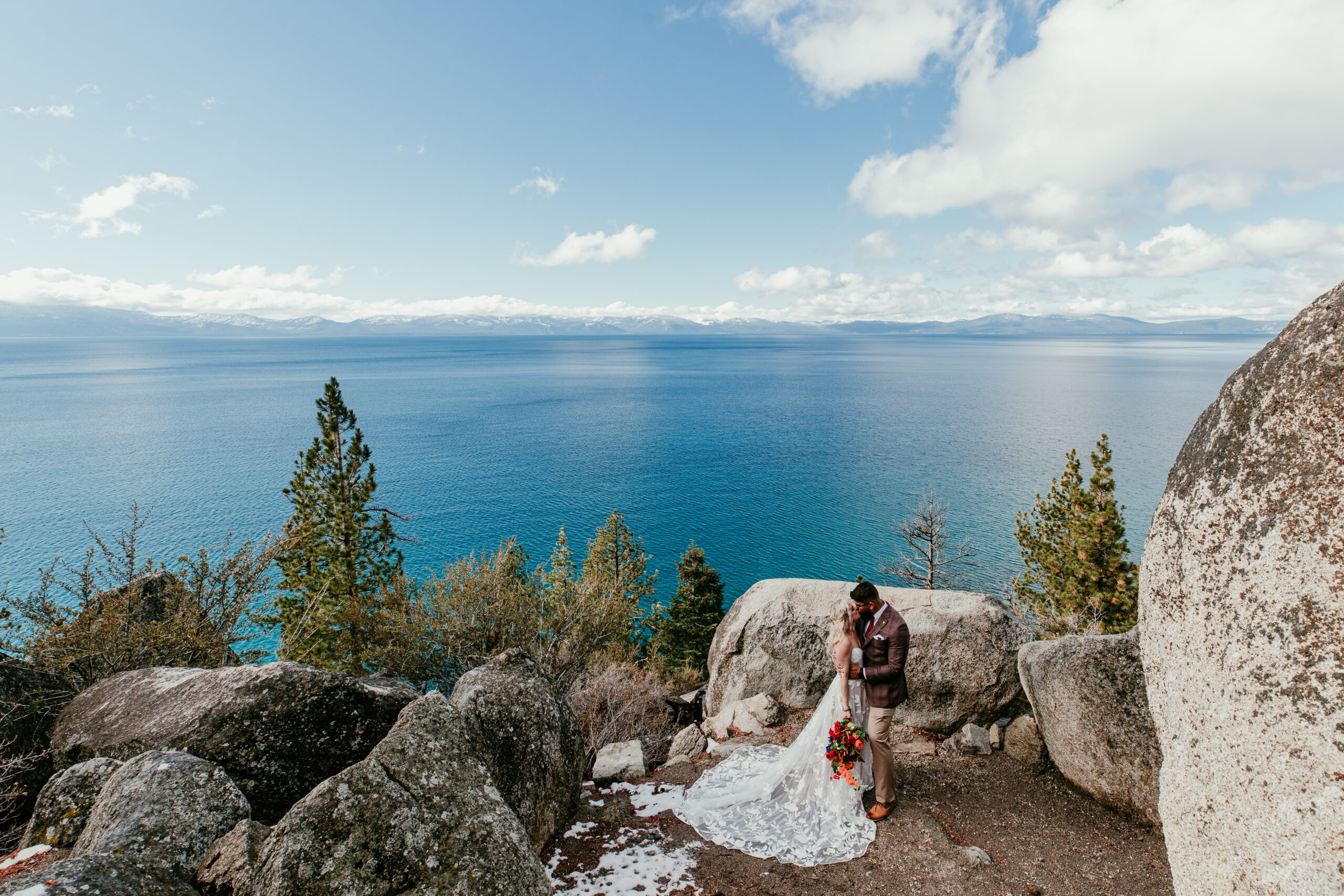 Couple embracing during a Lake Tahoe winter elopement at Logan Shoals Vista Point overlooking bright blue water and snow-covered mountains.