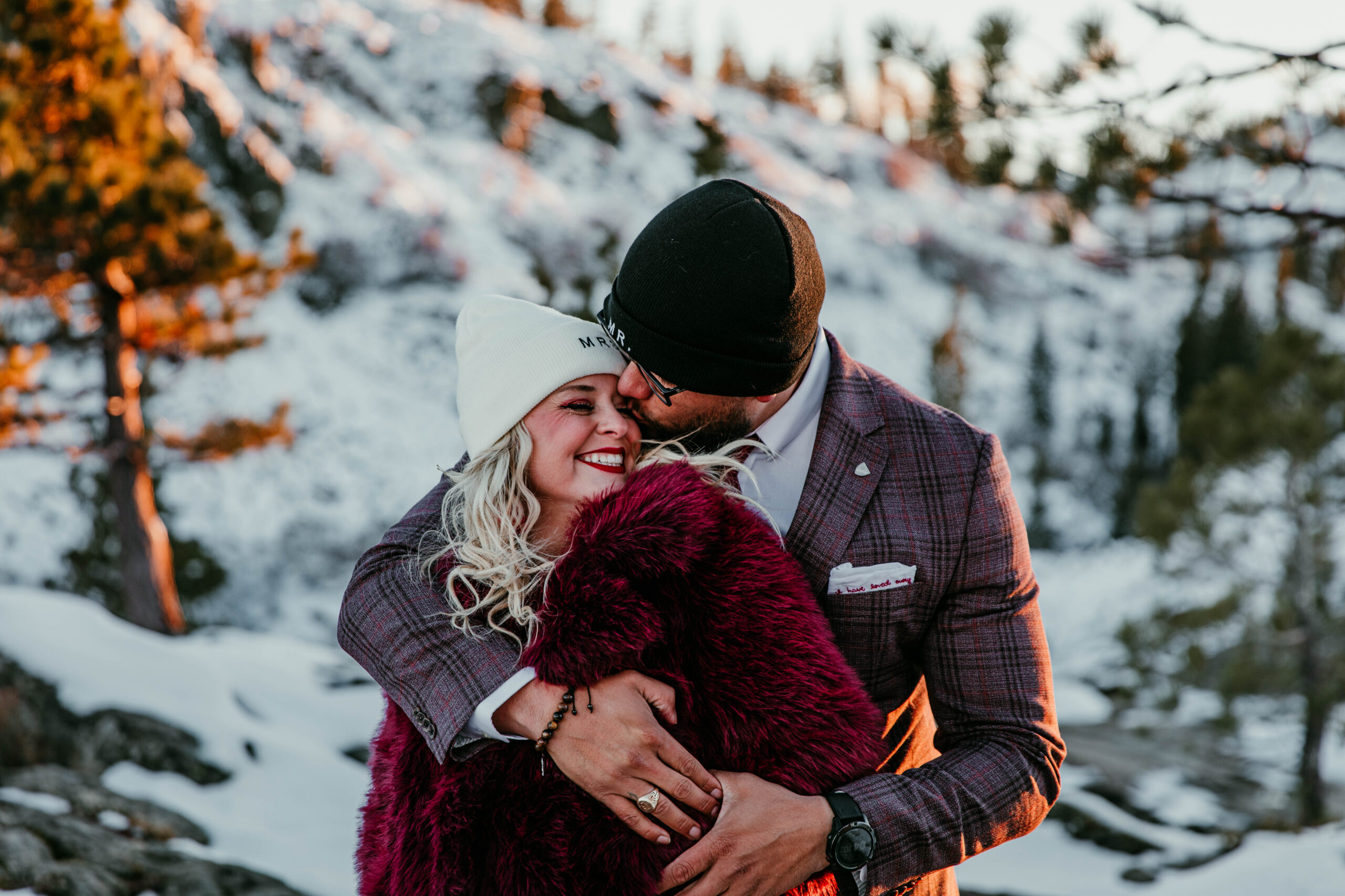 Windy winter portrait of bride and groom embracing at Donner Summit with snow-covered peaks behind them.