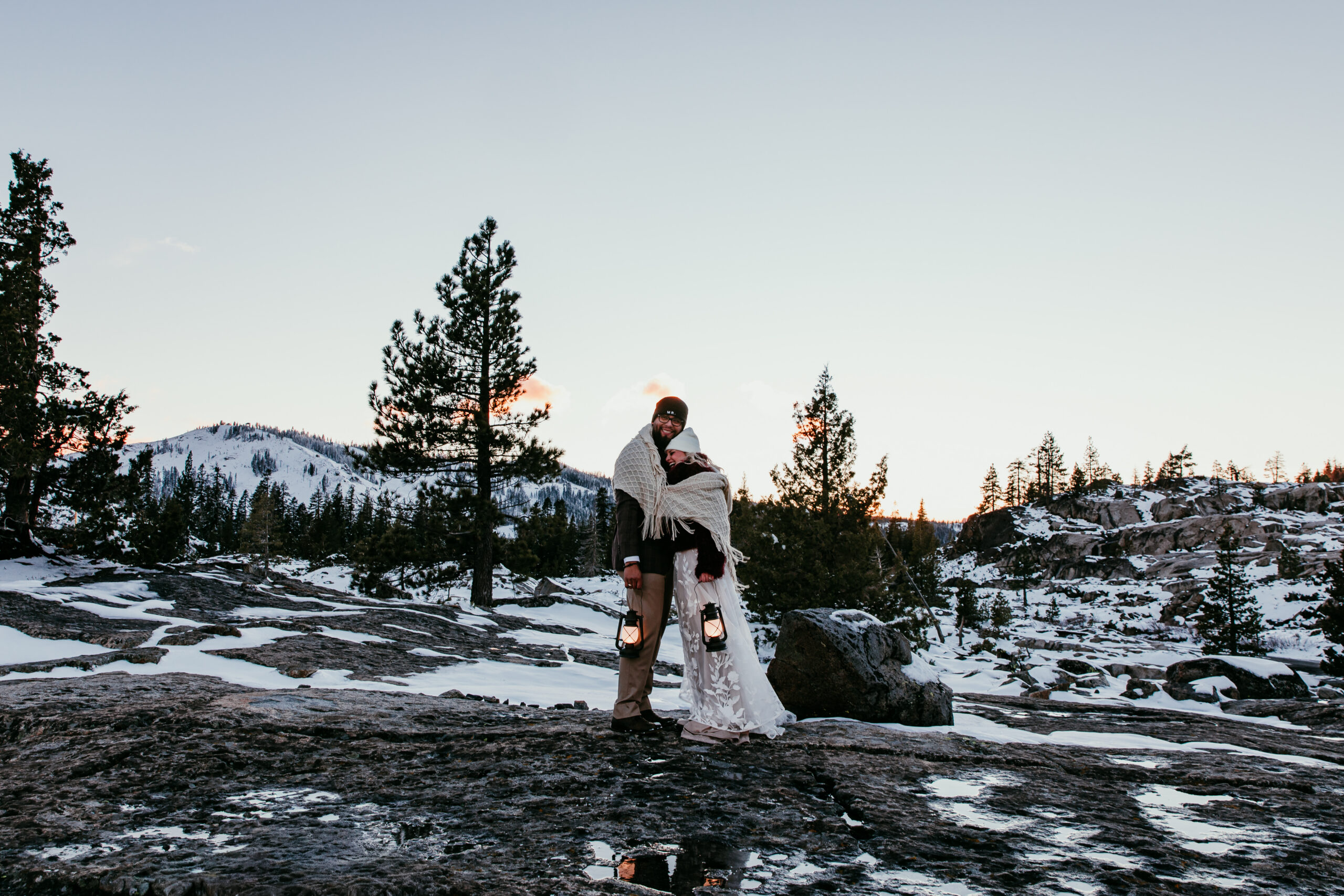 Windy winter portrait of bride and groom embracing at Donner Summit with snow-covered peaks behind them.