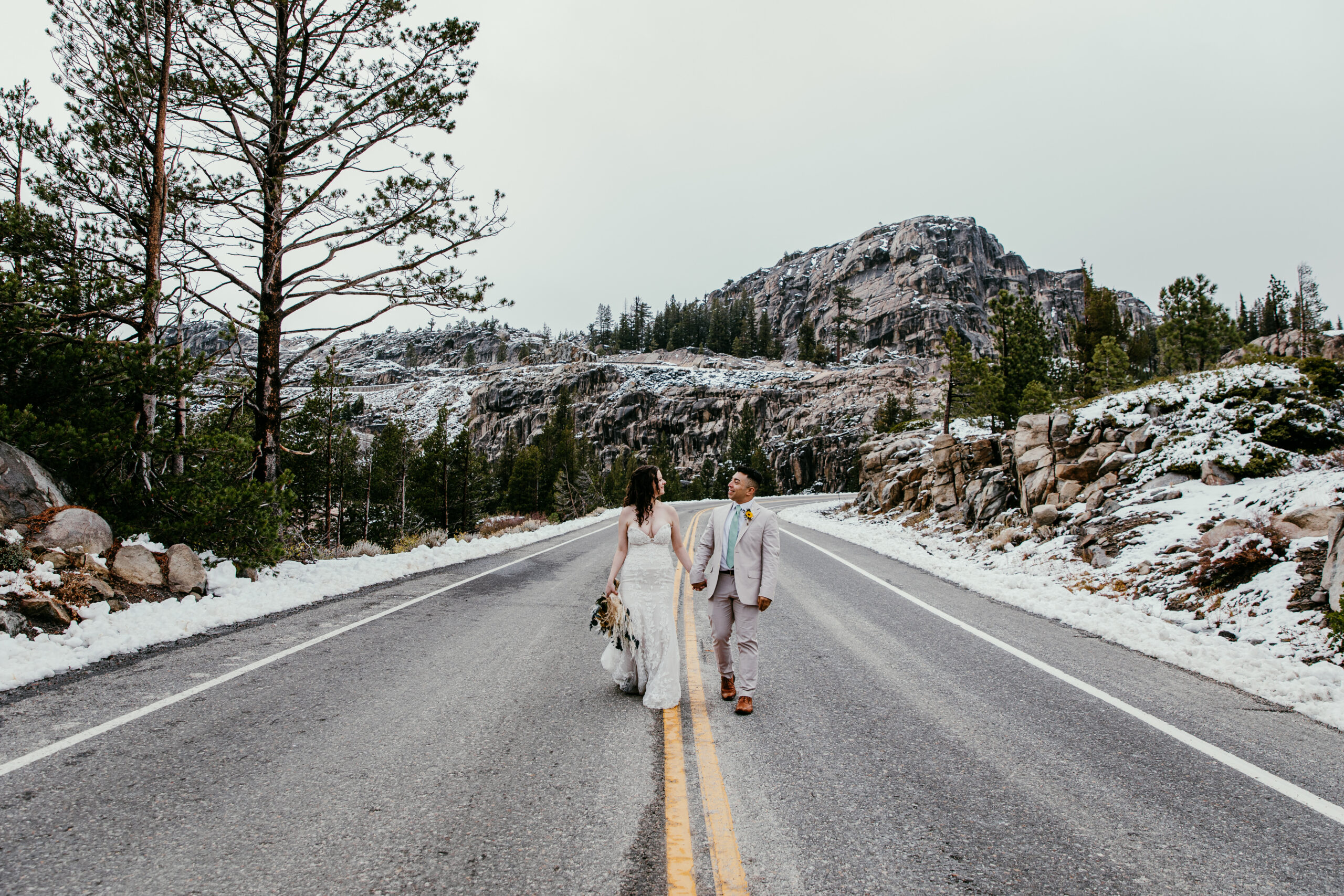 Newlyweds walking down a mountain road near Donner Summit with fresh snowfall along the roadside.