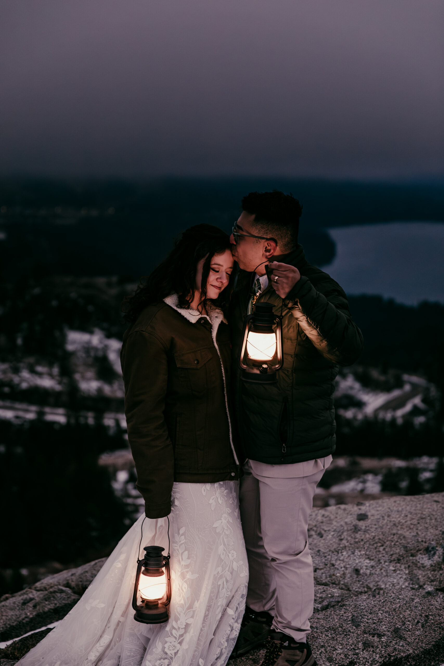 Bride and groom wrapped in jackets holding a lantern at blue hour on a snowy mountain overlook.