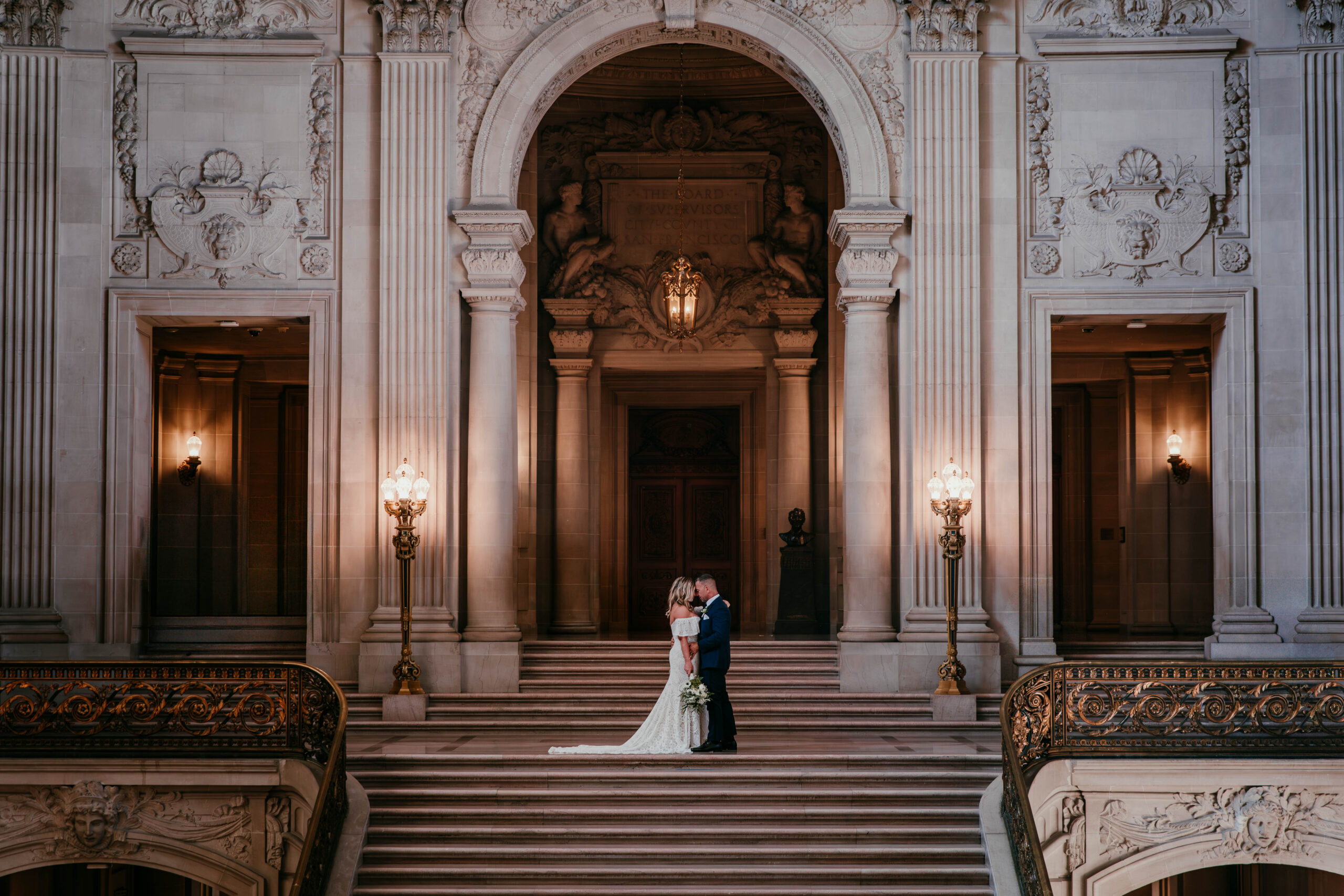 Wide angle wedding portrait on the grand staircase inside San Francisco City Hall with dramatic historic architecture.
