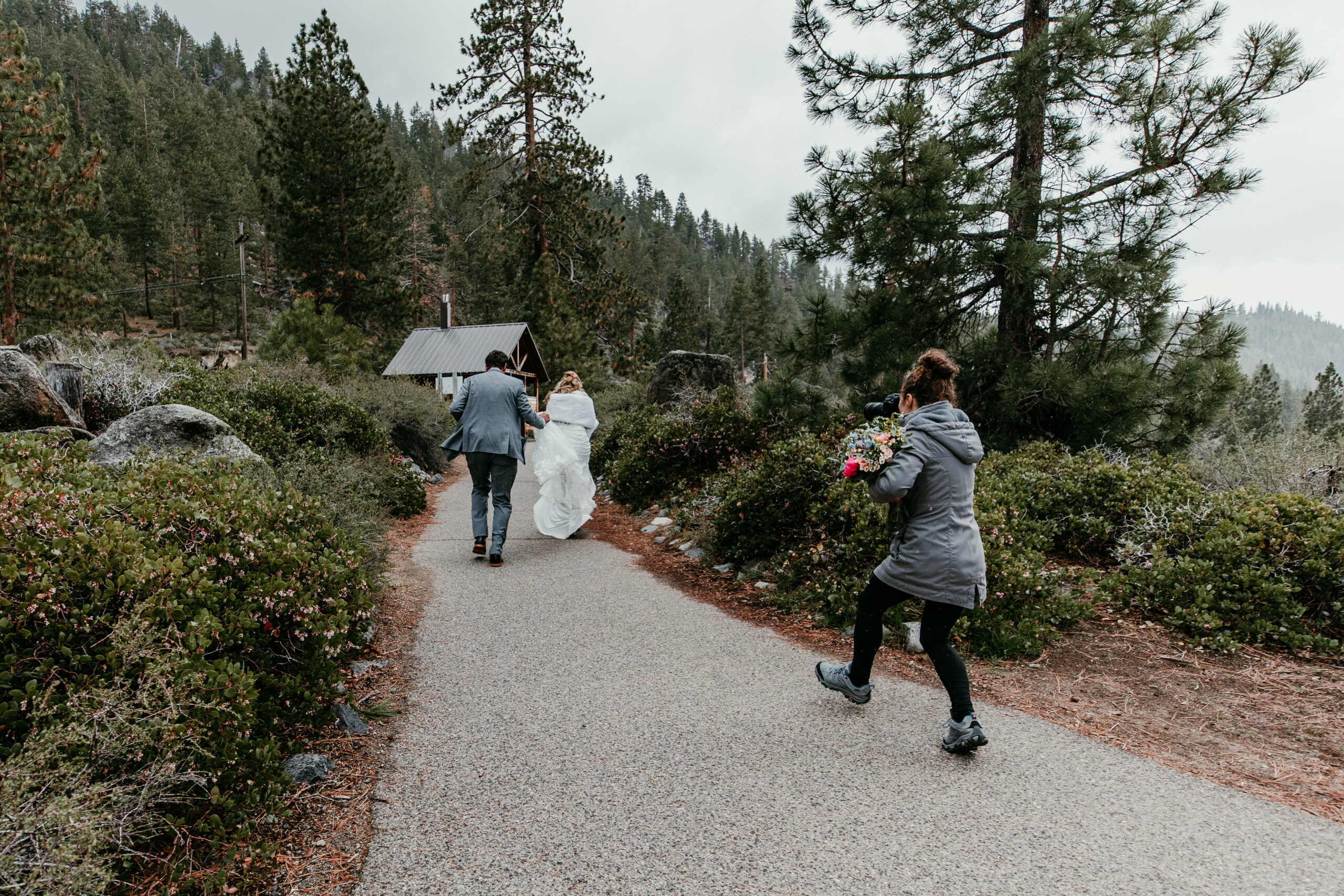 Bride and groom running hand in hand after their rainy Lake Tahoe elopement at Emerald Bay while their photographer follows behind capturing the moment.