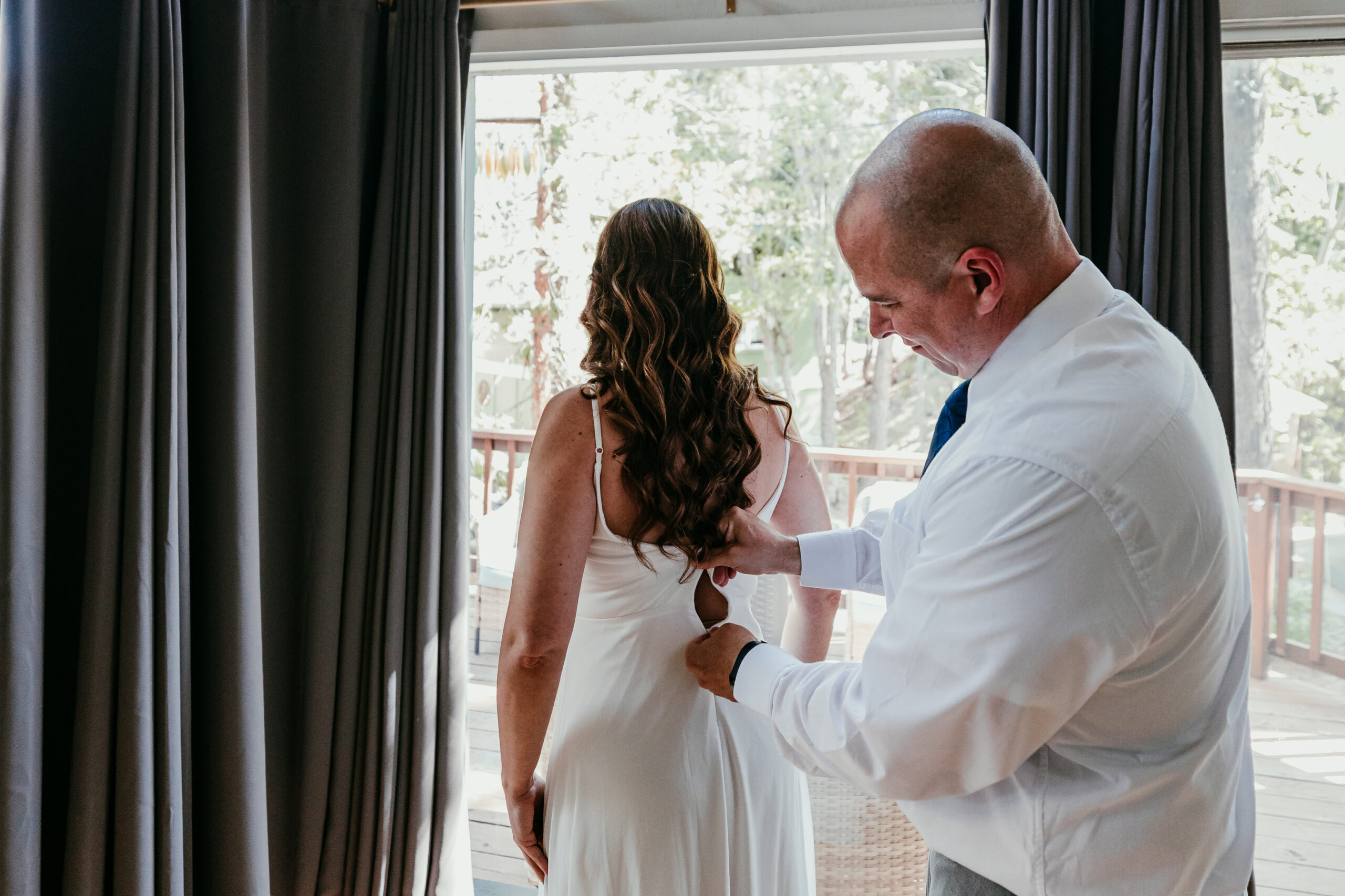 Groom fastening the back of his bride’s dress as they get ready together at their Lake Tahoe Airbnb before their Sand Harbor elopement.