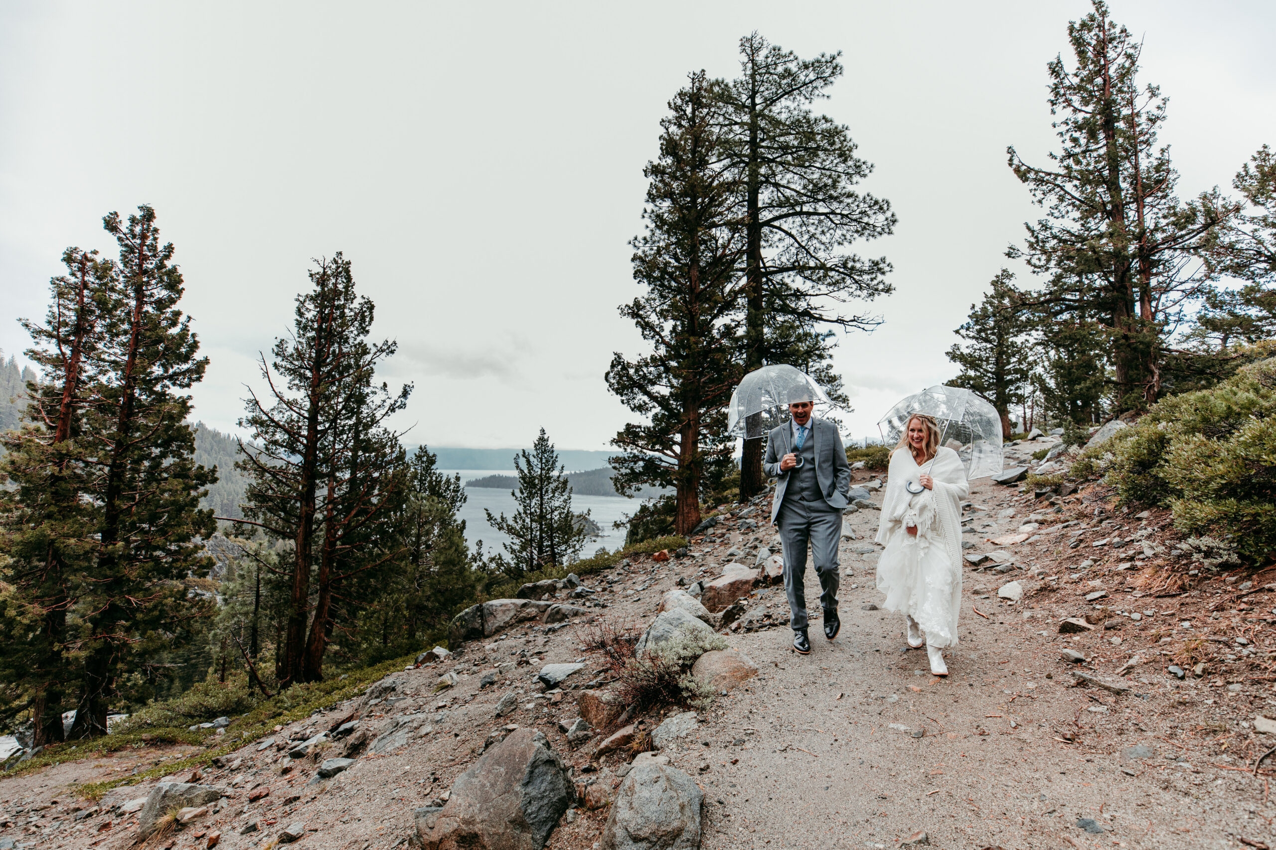 Bride and groom walking hand in hand with clear umbrellas during their rainy Lake Tahoe elopement on the trail to Eagle Falls at Emerald Bay.