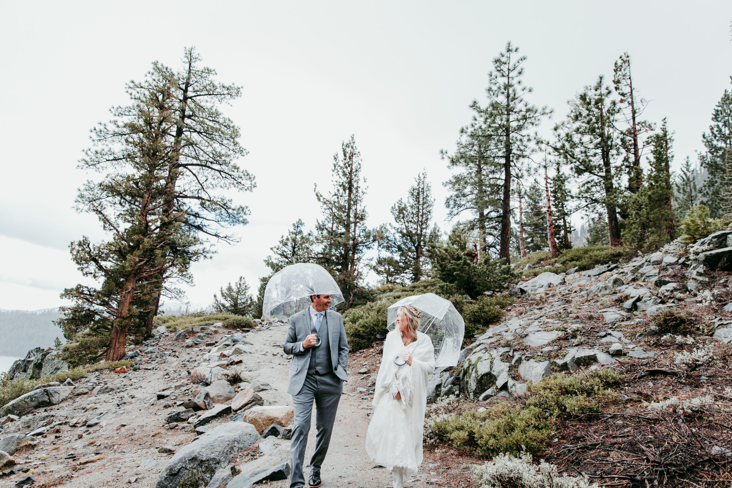 Bride and groom smiling at each other under clear umbrellas during their rainy Lake Tahoe elopement near Eagle Falls at Emerald Bay.
