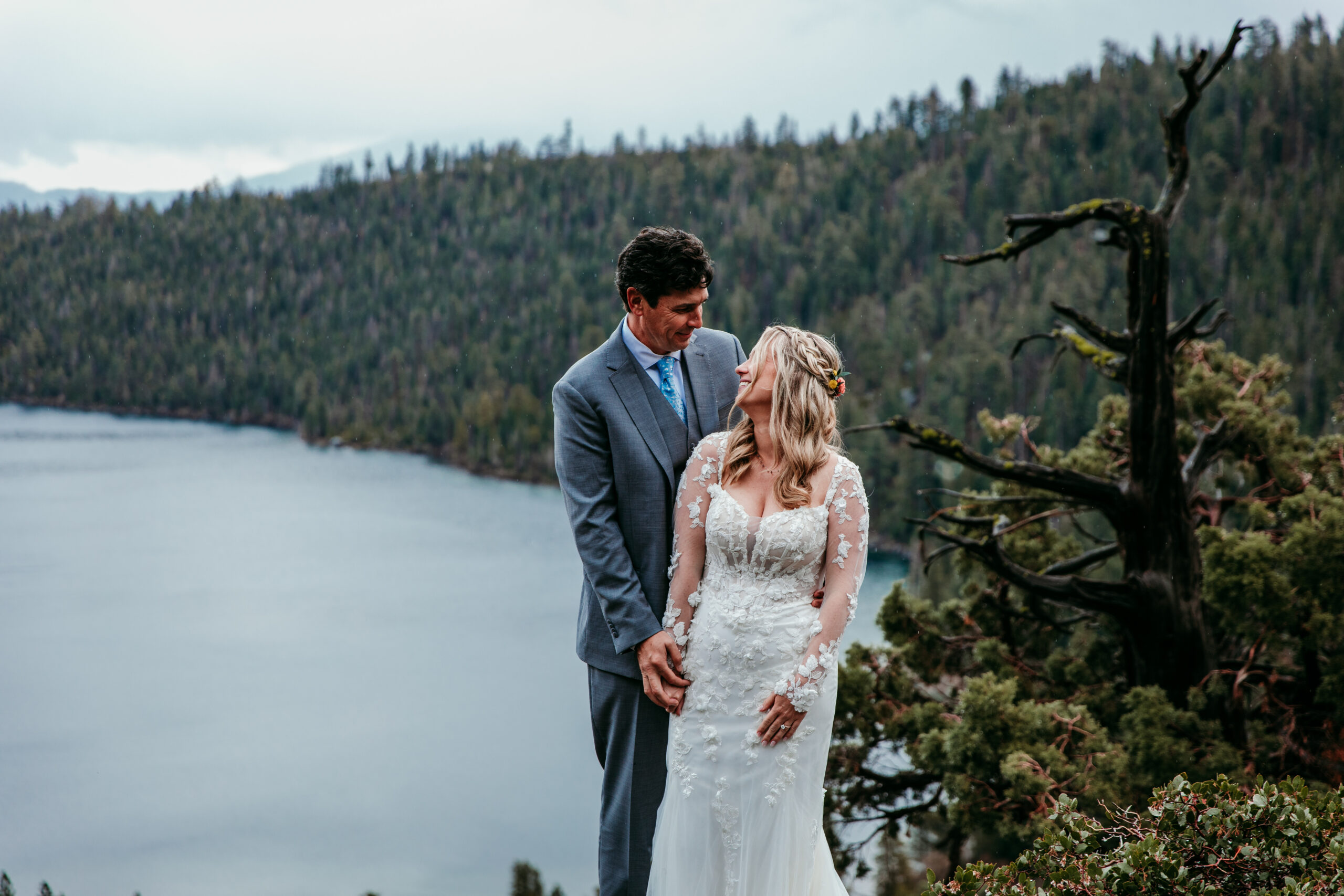 Bride and groom holding hands during their rainy Lake Tahoe elopement at Emerald Bay with the lake and forested shoreline in the background.
