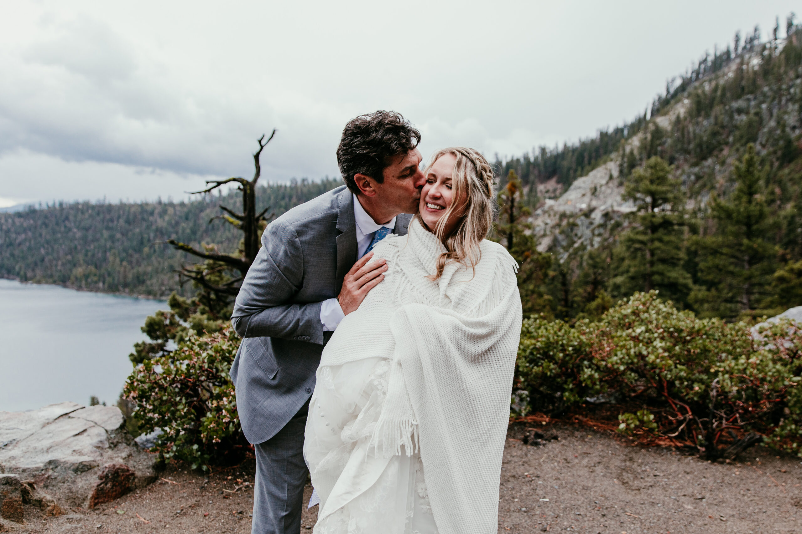 Groom kissing the bride's cheek as she wraps up in a blanket during their rainy Lake Tahoe elopement at Emerald Bay.
