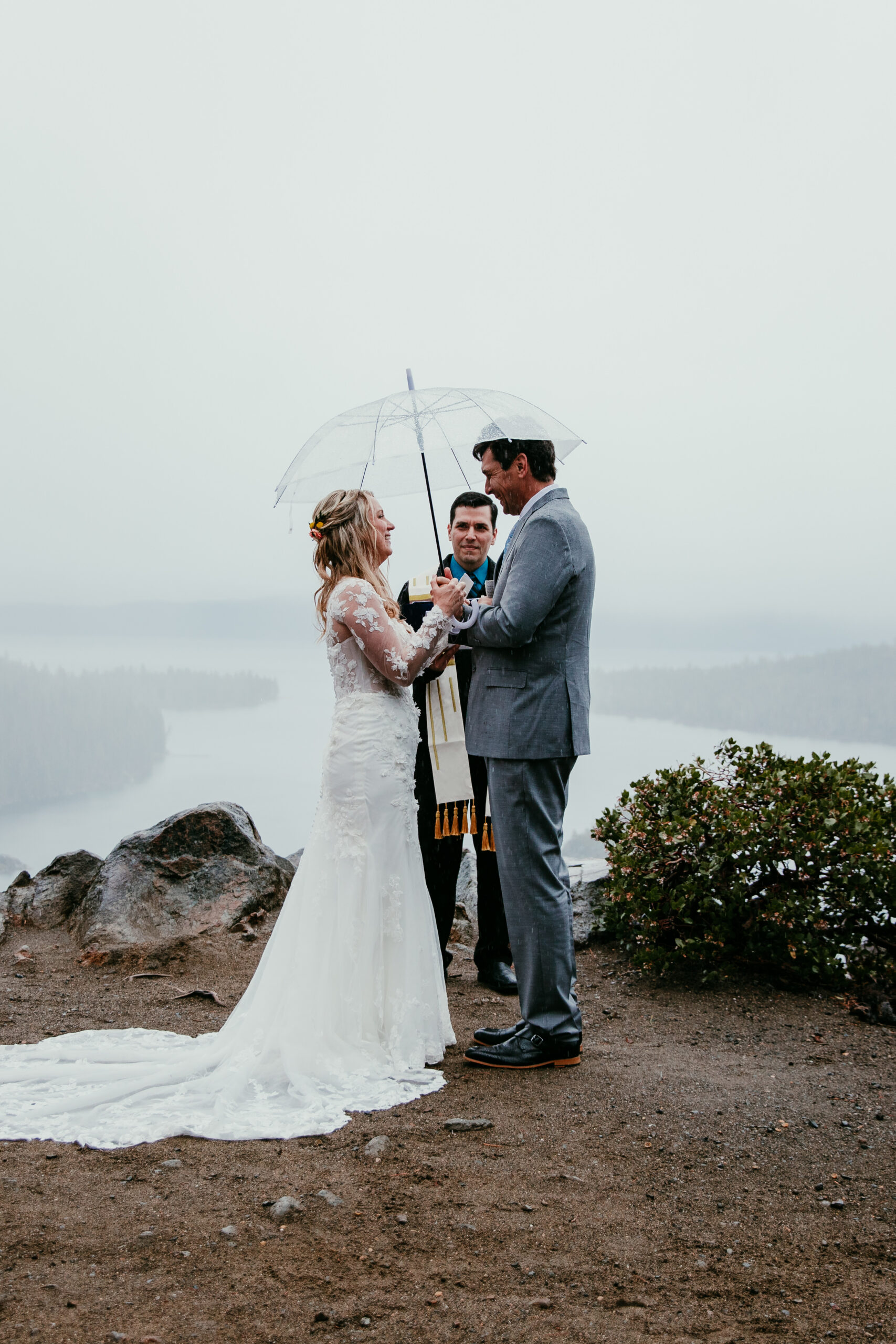 Bride and groom exchanging vows under a clear umbrella during a rainy Lake Tahoe elopement ceremony at Emerald Bay with fog covering the lake.