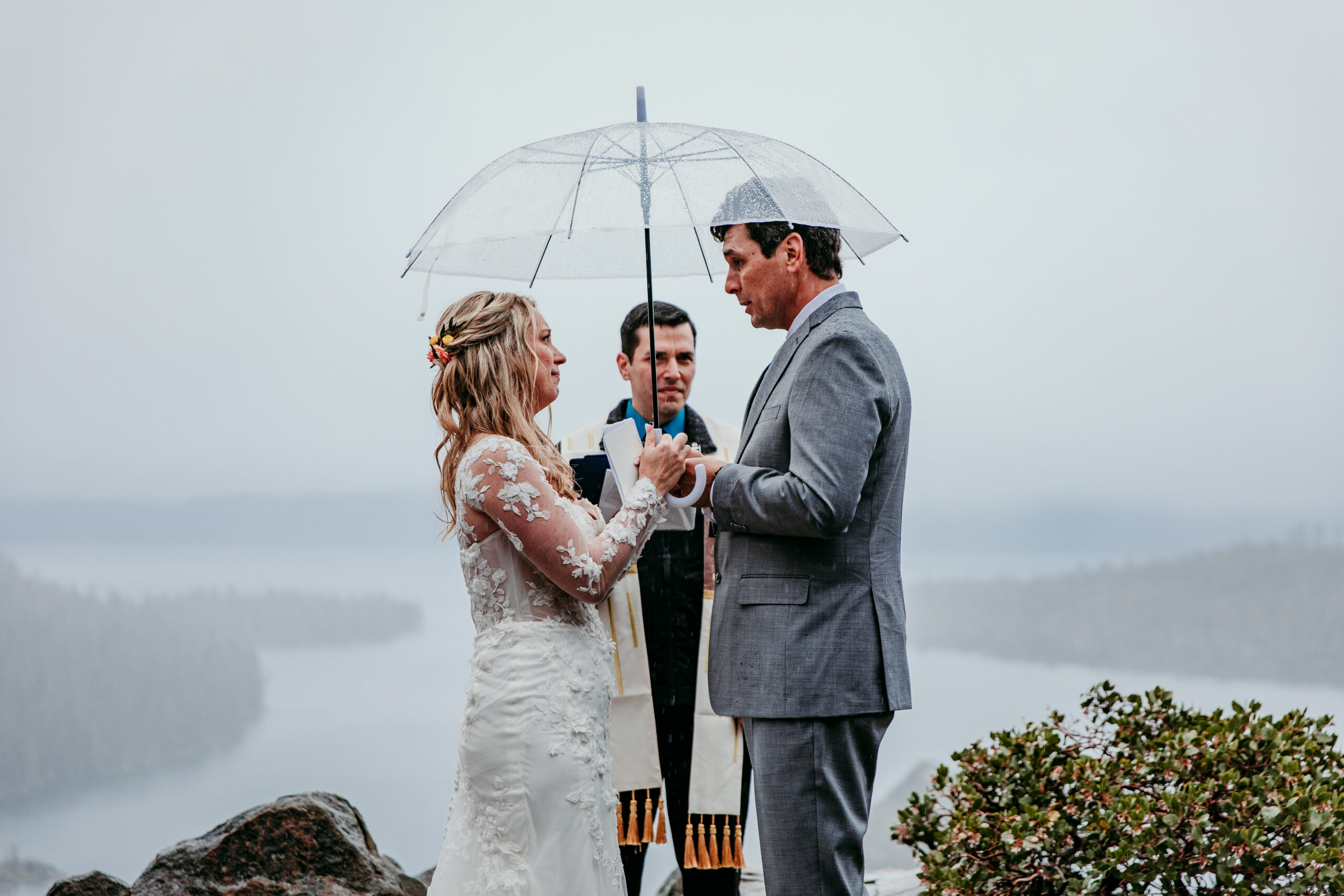 Bride and groom reading their vows under a clear umbrella during their rainy Lake Tahoe elopement ceremony at Emerald Bay with fog over the lake.