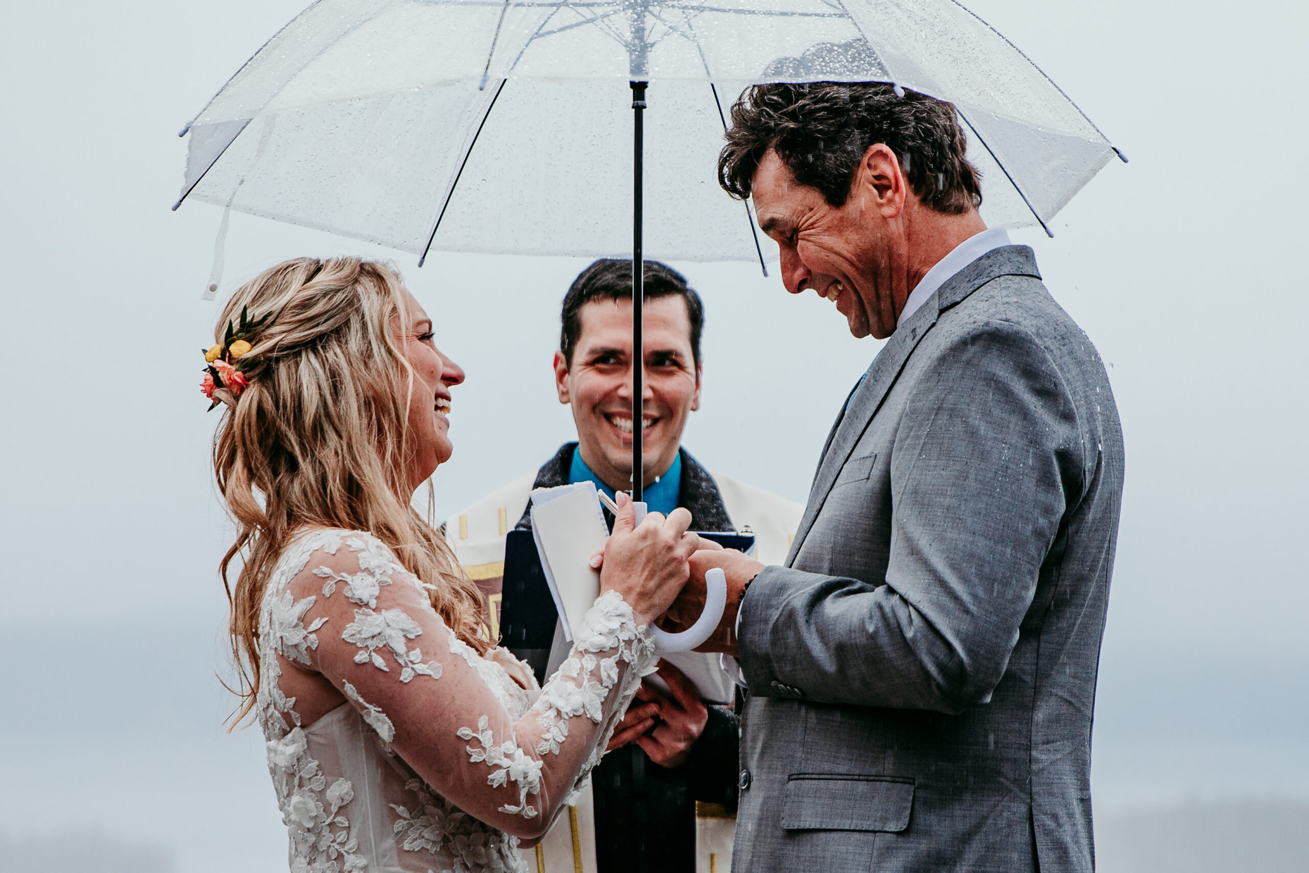 Bride and groom smiling as they exchange rings under a clear umbrella during their rainy Lake Tahoe elopement ceremony at Emerald Bay.