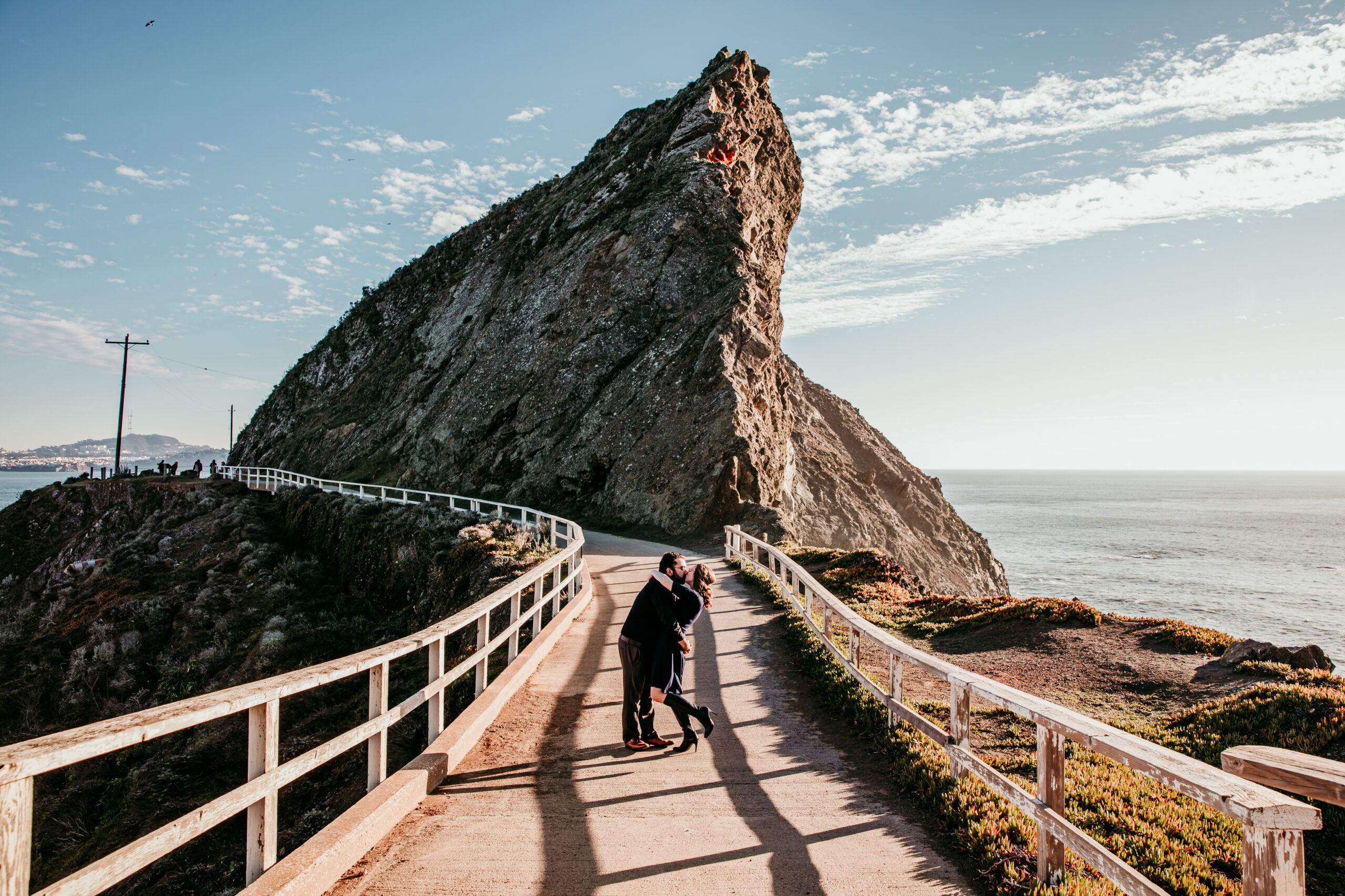 Couple kissing on a coastal path beside a dramatic rock formation overlooking the Pacific Ocean.