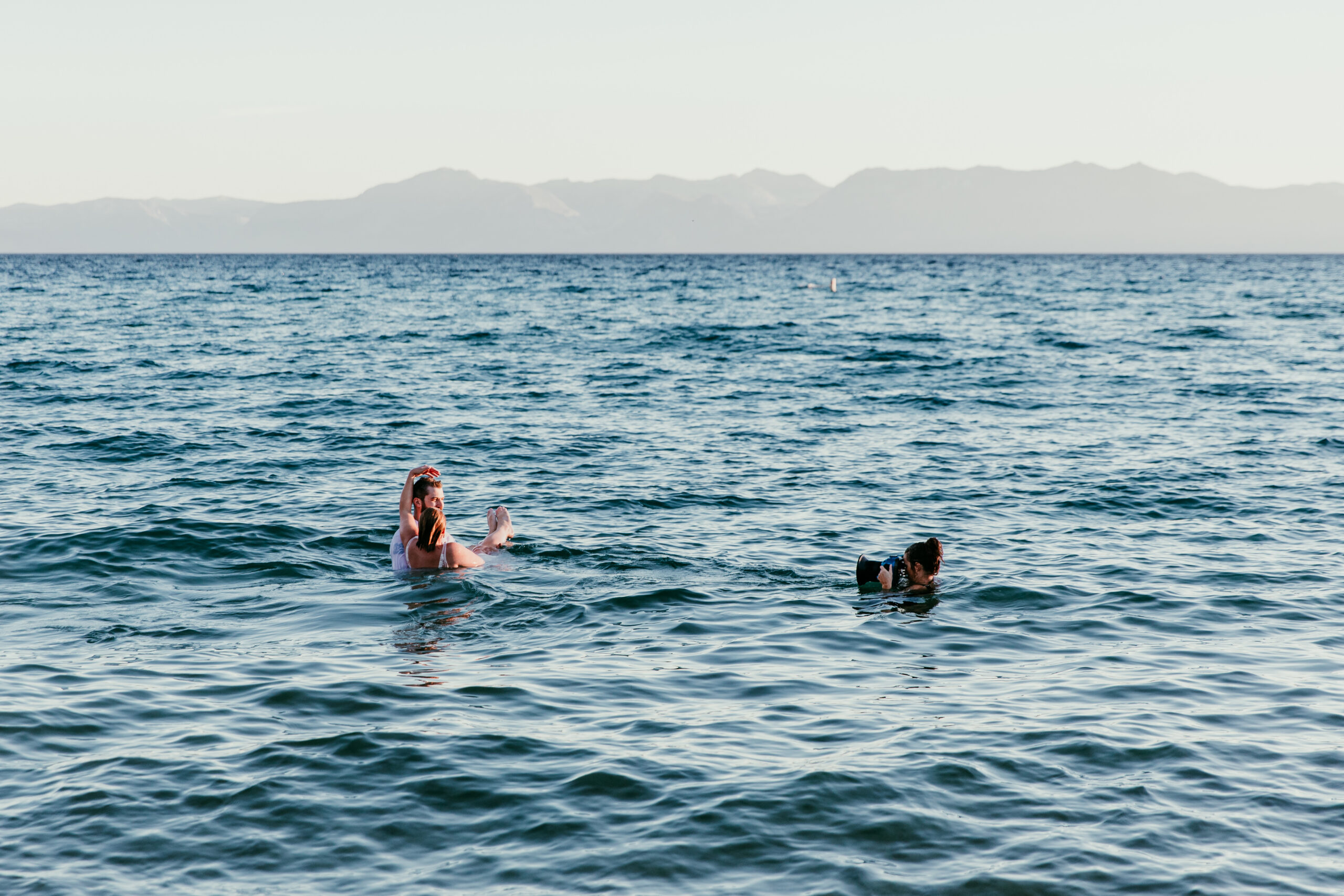 Photographer using a water housing to photograph a couple embracing in Lake Tahoe, with blue waves and distant mountains behind them.