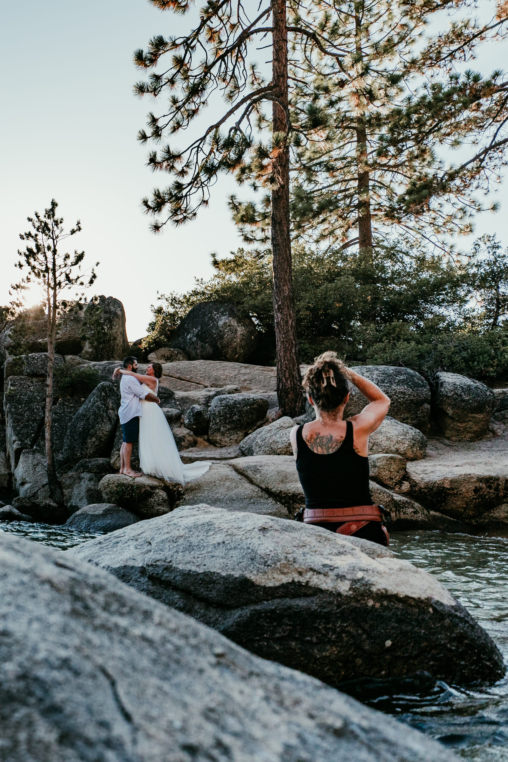 Photographer capturing SJ and Dave embracing on granite boulders at Sand Harbor, surrounded by pine trees.
