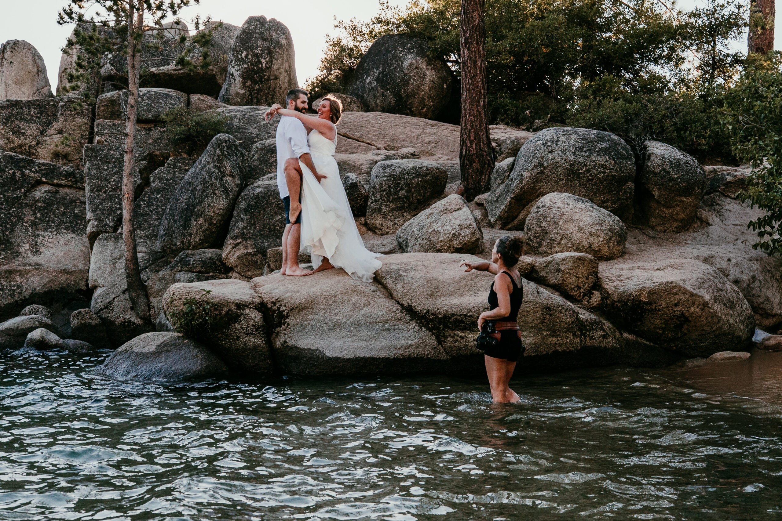Photographer standing in shallow water to photograph SJ and Dave kissing on the rocks beside Lake Tahoe.