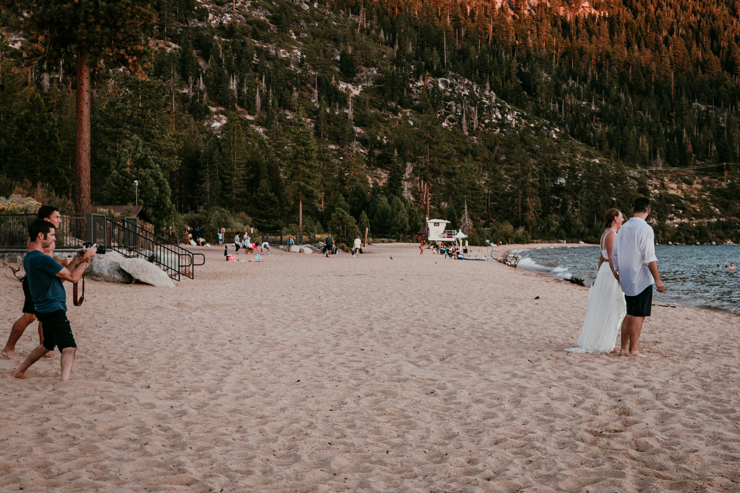 Videographer and photographer filming SJ and Dave together on the sandy beach at Sand Harbor, with a forested hillside behind them.