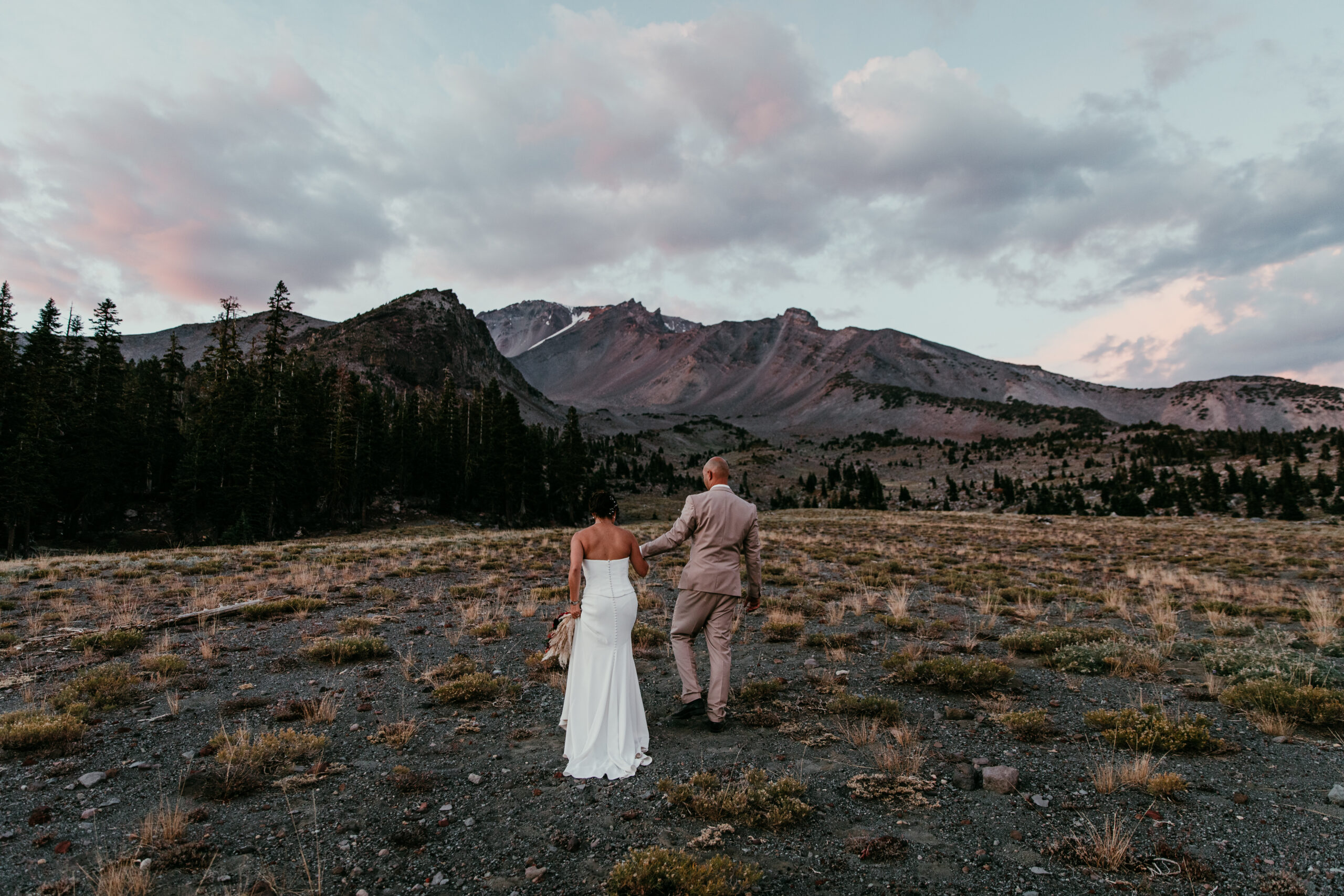 Newlyweds holding hands and walking toward a rugged mountain ridge beneath a cloudy evening sky.
