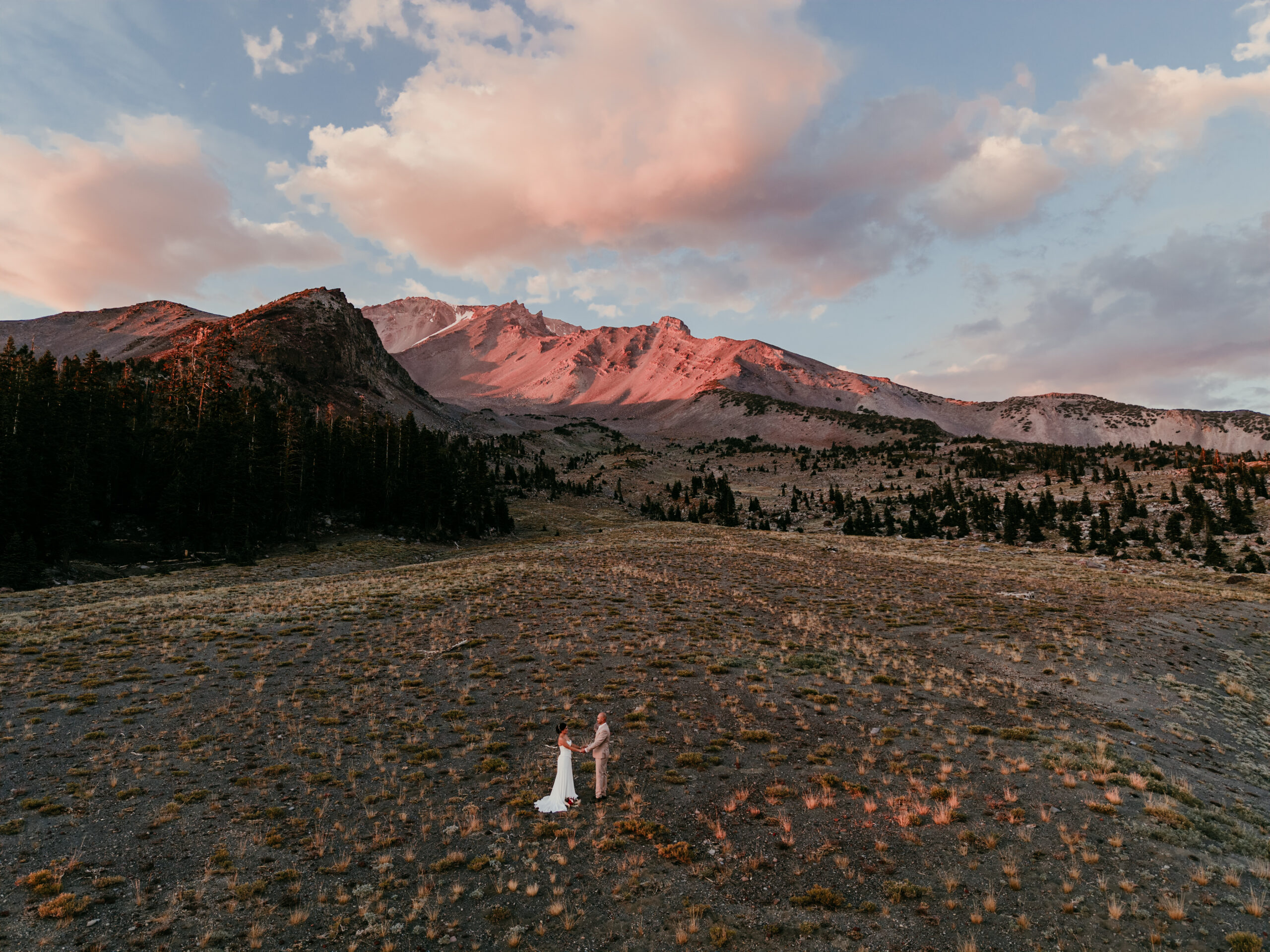 Couple holding hands in an open meadow beneath Mount Shasta, with pink sunset light on the rocky slopes and clouds overhead.