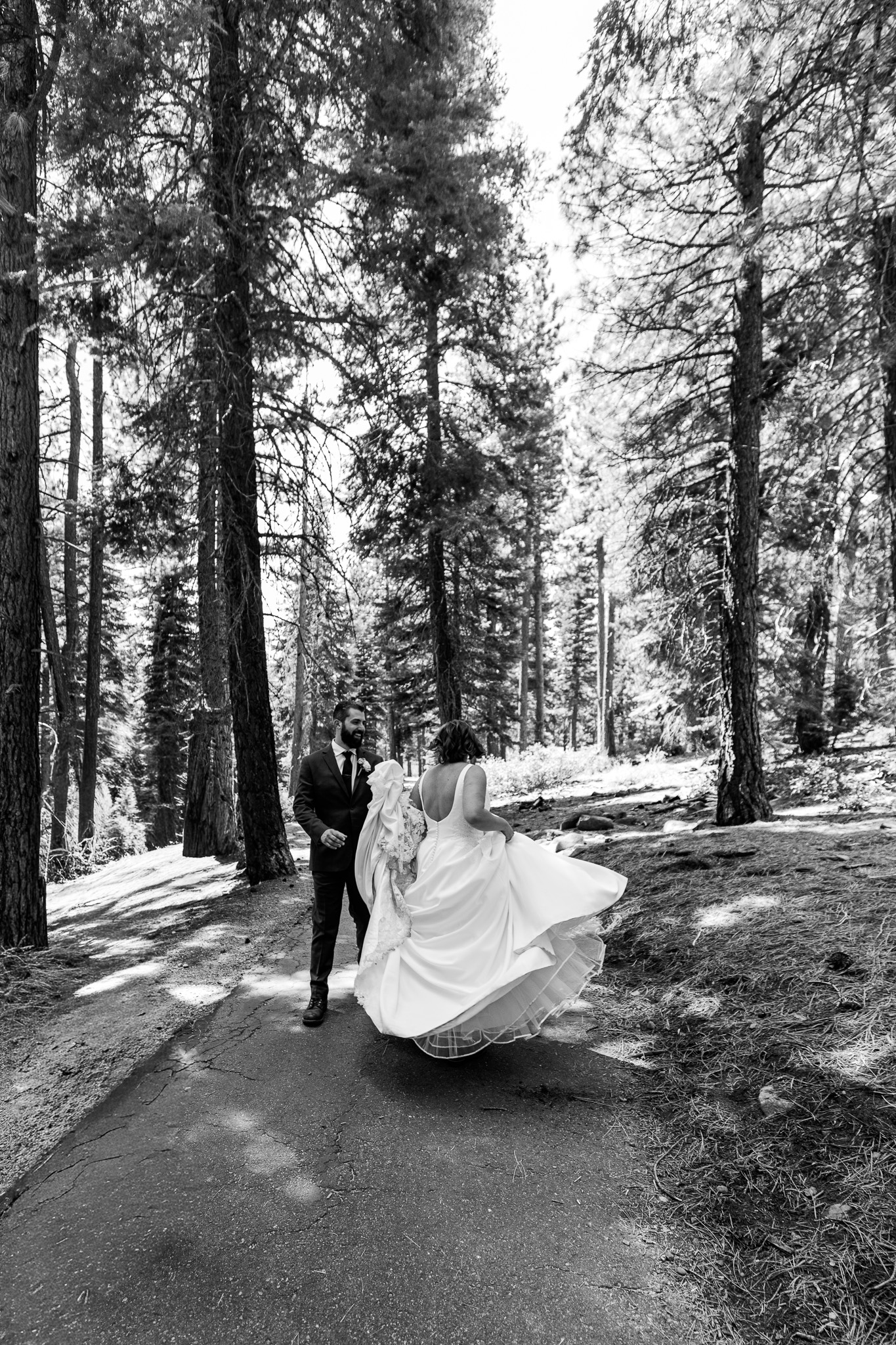 Black-and-white photo of newlyweds walking through the Tahoe forest as the bride’s wedding dress swings around her.