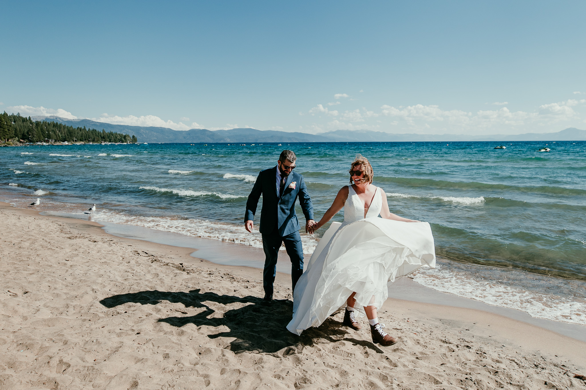 Newlyweds holding hands on the beach, wearing sunglasses beside Lake Tahoe’s blue water.