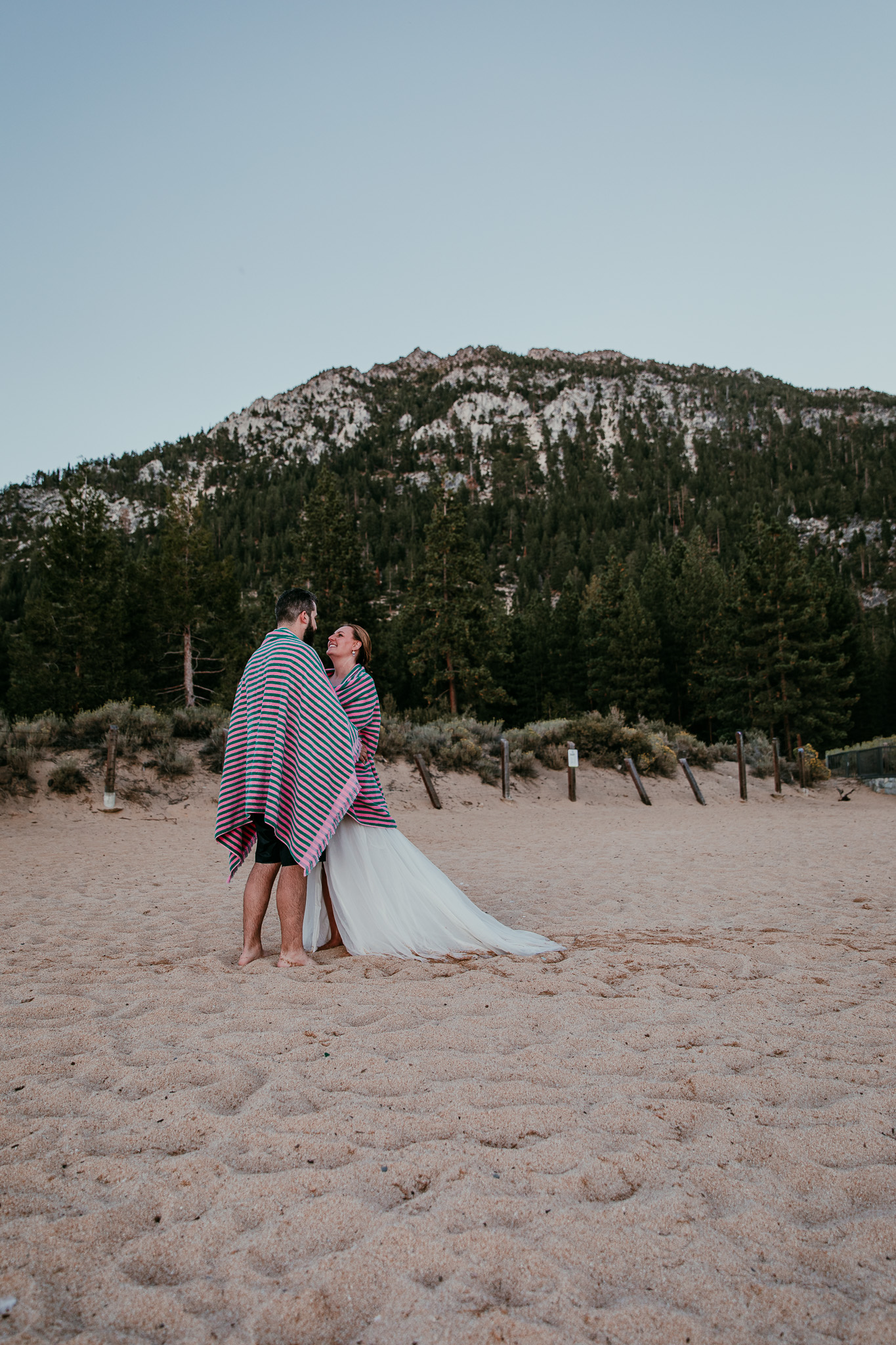 SJ and Dave cuddling on the beach beneath a striped towel, with a rocky, forested mountain behind them.