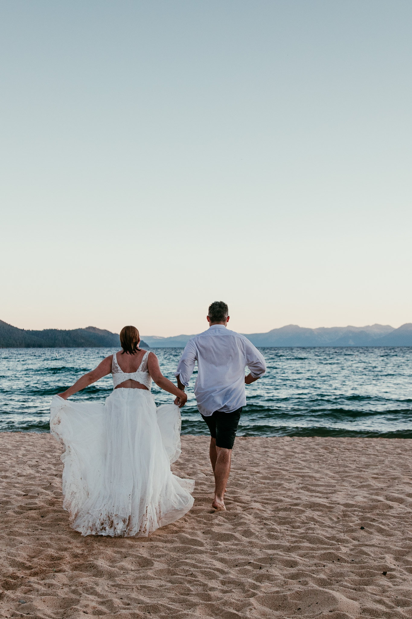 Newlyweds walking hand in hand toward Lake Tahoe, with SJ lifting her wedding dress above the sand.