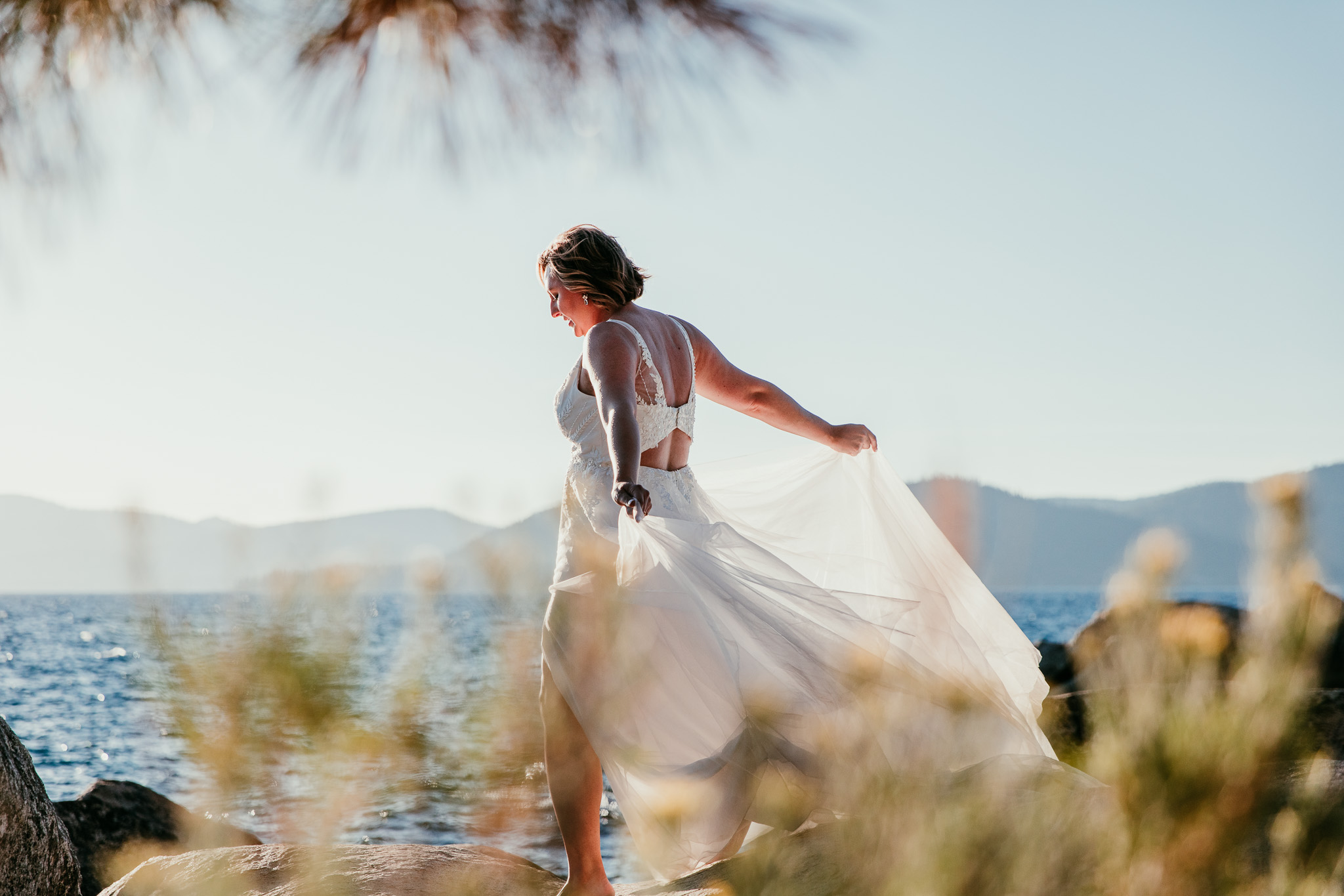 SJ holding out her wedding dress skirt beside Lake Tahoe, photographed through softly blurred shoreline plants.