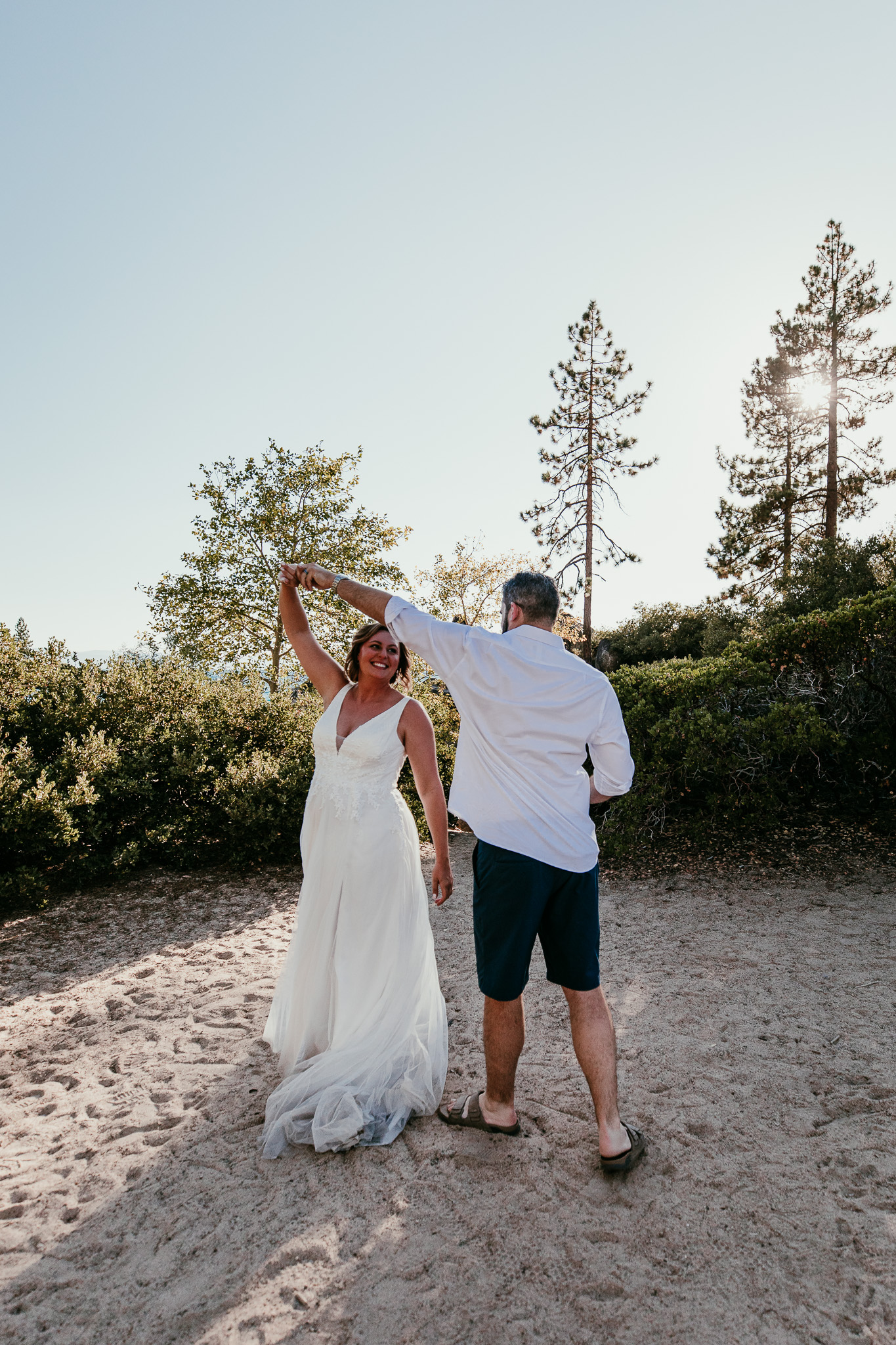 Dave twirling SJ on the sand, wearing a white shirt and shorts beside her flowing wedding dress.