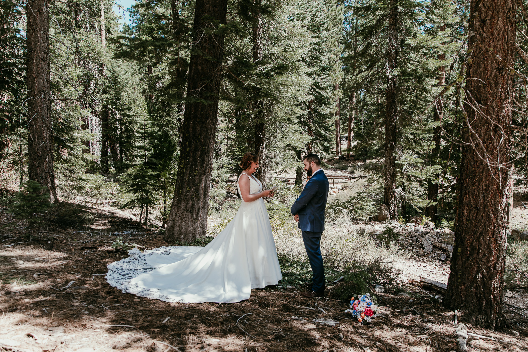 Bride and groom exchanging vows among Lake Tahoe pine trees, with her wedding dress train spread across the forest floor.