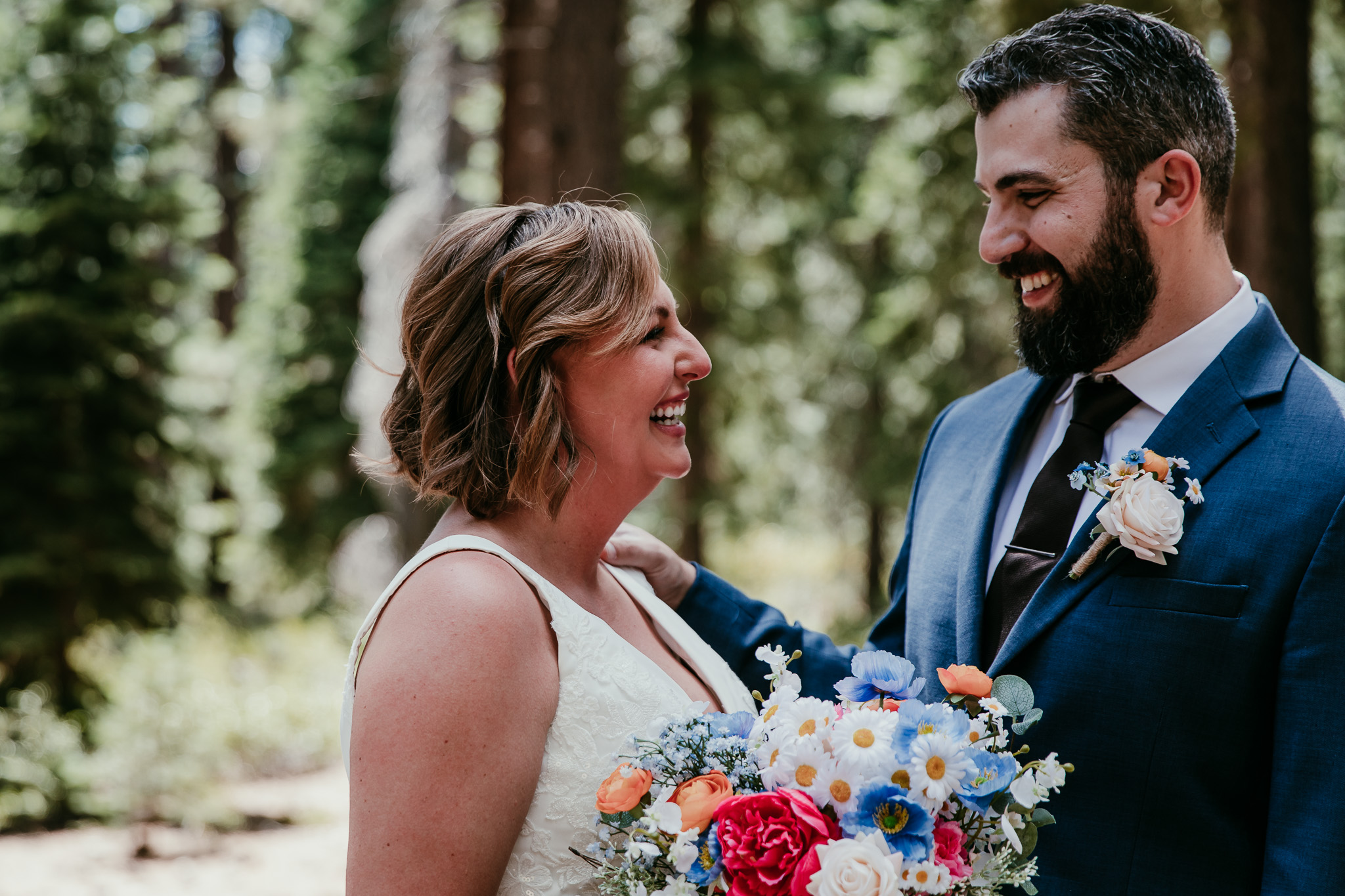 Bride and groom laughing together in the forest, with her colorful bouquet and his blue suit.