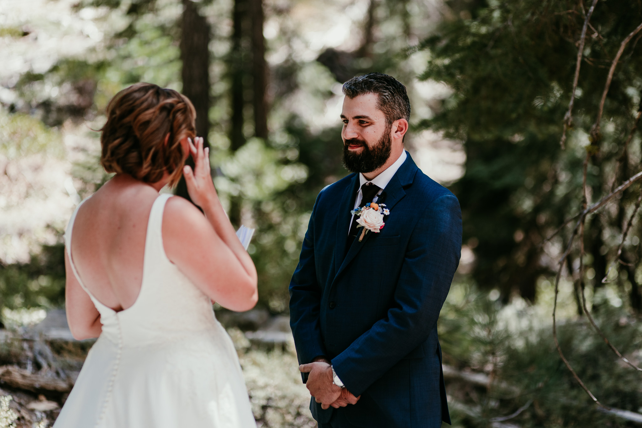 Groom in a blue suit smiling at his bride as she holds her vow book during their forest ceremony.