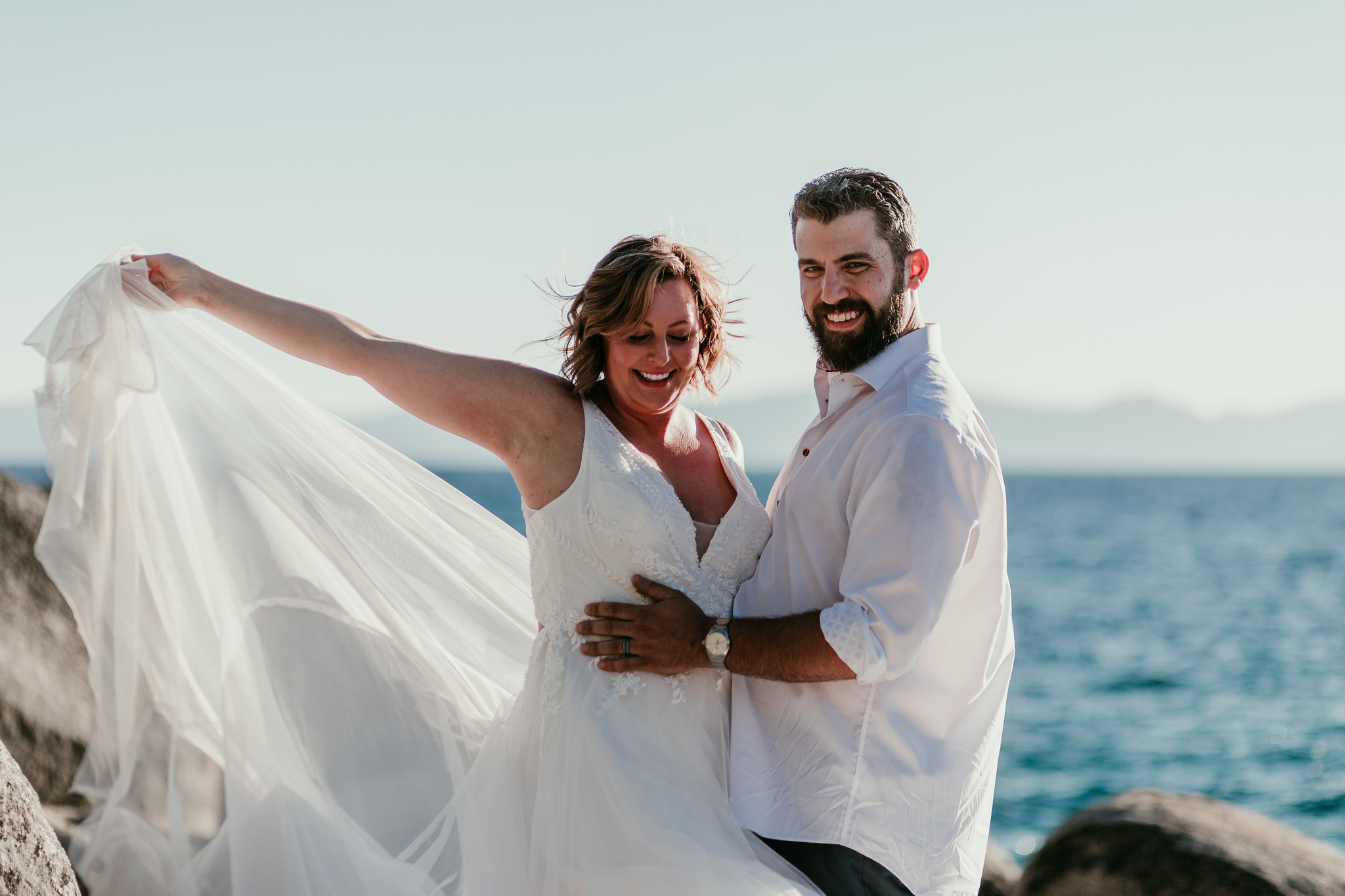 SJ lifting her flowing wedding dress skirt while Dave holds her waist, both smiling beside Lake Tahoe.