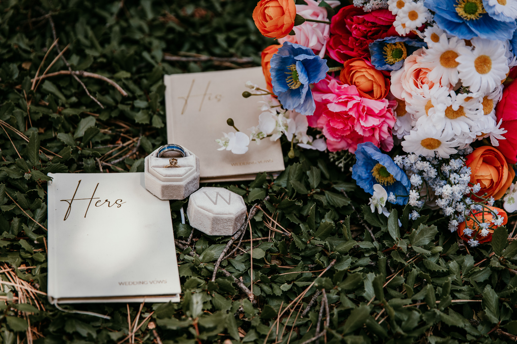 Wedding rings and vow books beside a colorful bouquet on the forest floor during a summer Lake Tahoe elopement.