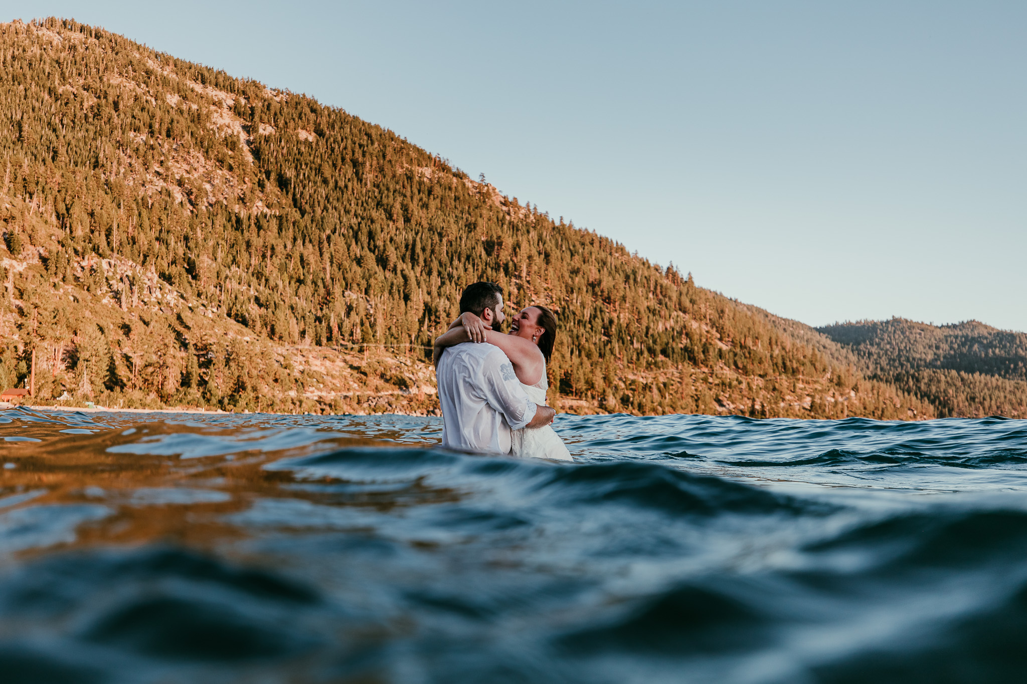SJ and Dave embracing in Lake Tahoe, photographed at water level with golden mountains behind them.